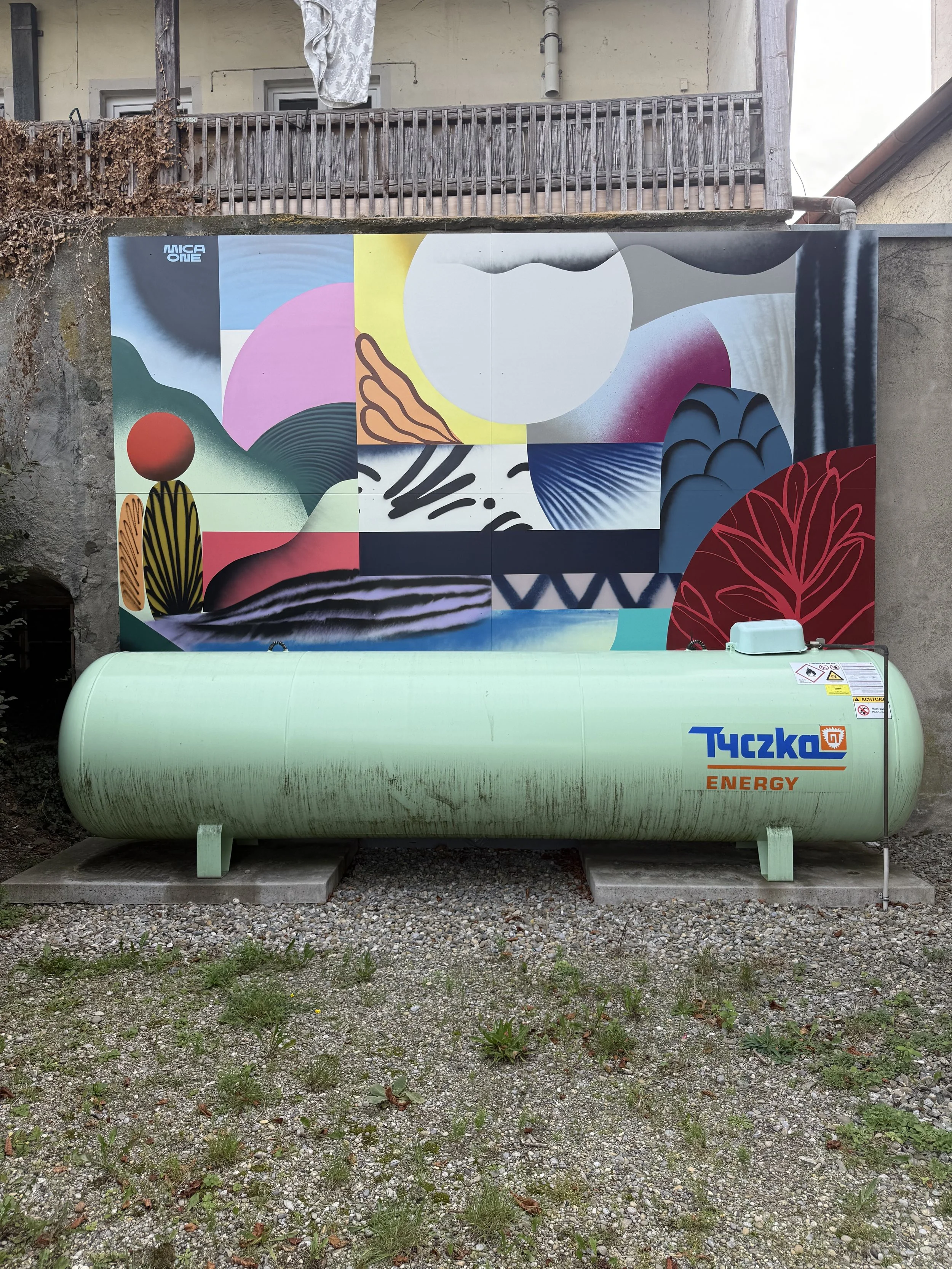 Ein grüner Energie-Tank mit dem Aufkleber 'Tyczko ENERGY' steht auf einer gepflasterten Fläche mit Gras und kleinen Pflanzen. Im Hintergrund ist eine Wand mit einem bunten Wandbild und einem Balkon sichtbar.