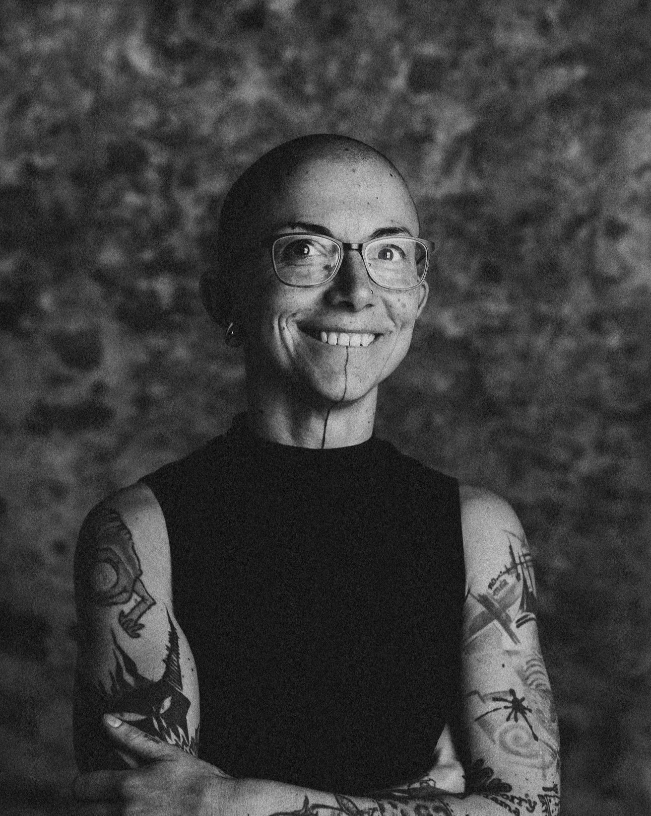 Schwarz-Weiß-Fotografie einer Tattoo-tragenden Frau mit  Lächeln vor einem Steinmauer-Hintergrund.