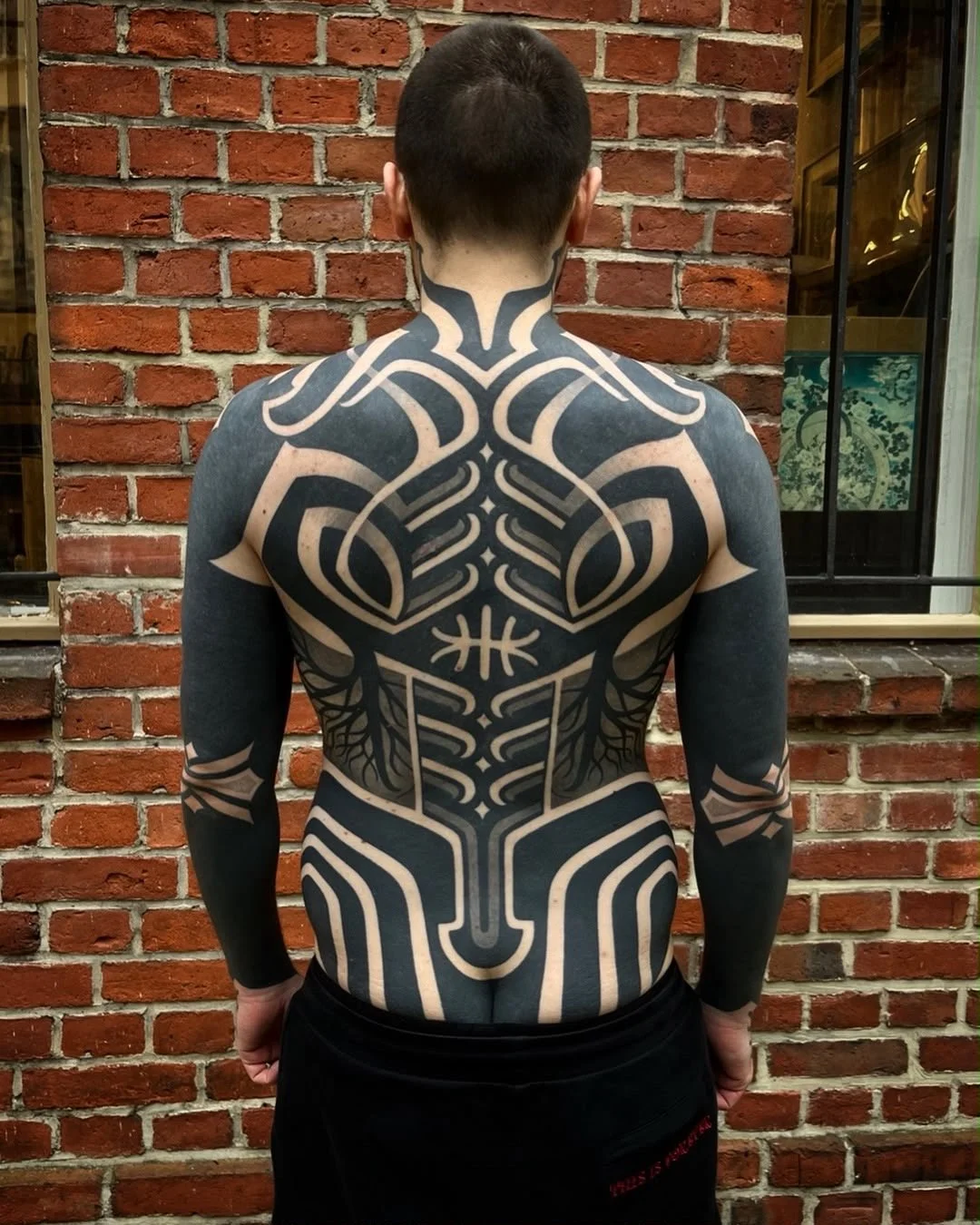 Rückansicht einer Person mit großflächigem schwarzen, geometrischen Tattoo über Rücken, Schultern und Arme vor einer Backsteinwand.