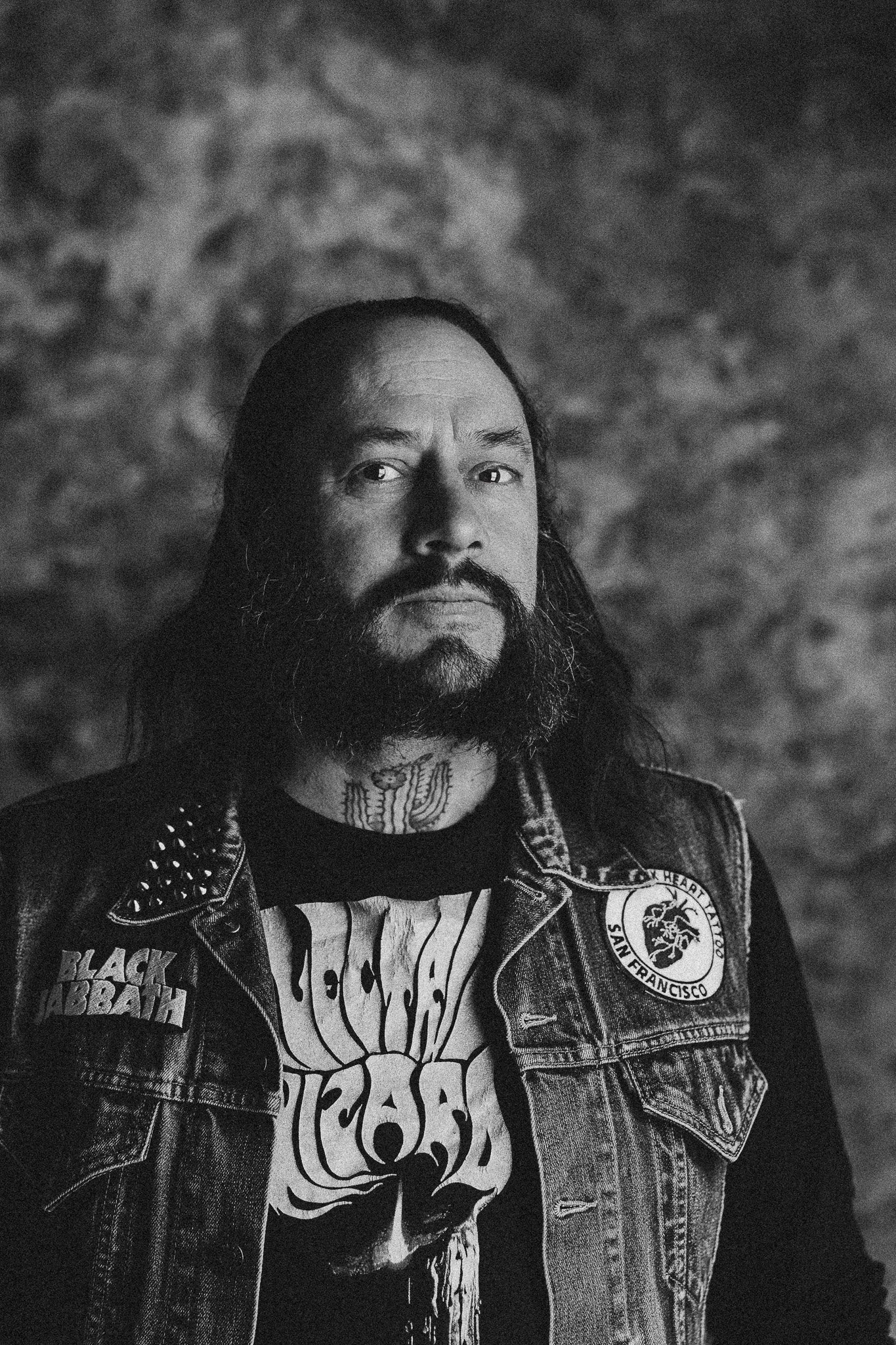 Schwarz-weiß-Foto eines Mannes mit langen Haaren, Vollbart und Tattoos, trägt ein Skull-T-Shirt und eine Denim-Jacke, vor einem unscharfen, grauen Hintergrund.
