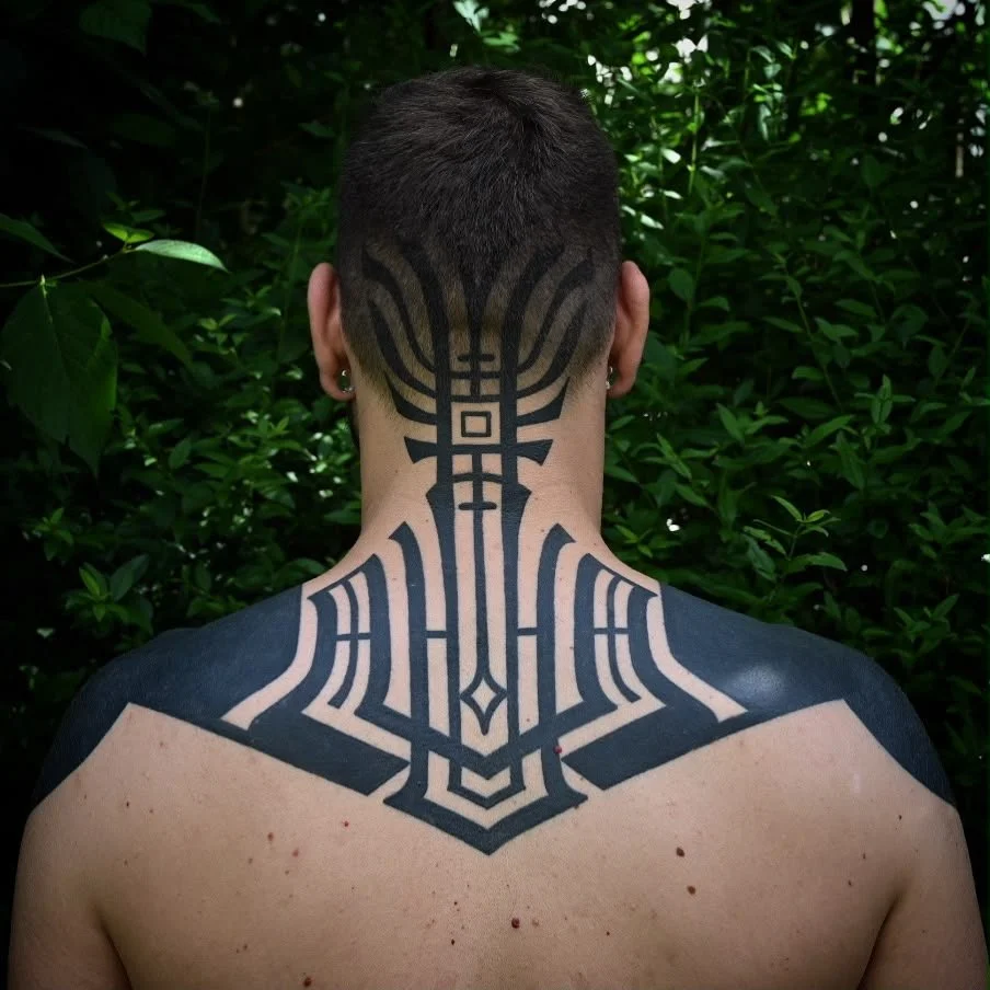 Rückansicht einer Person mit großem geometrischem schwarzem Tattoo am Nacken und oberen Rücken vor grünem Laub.