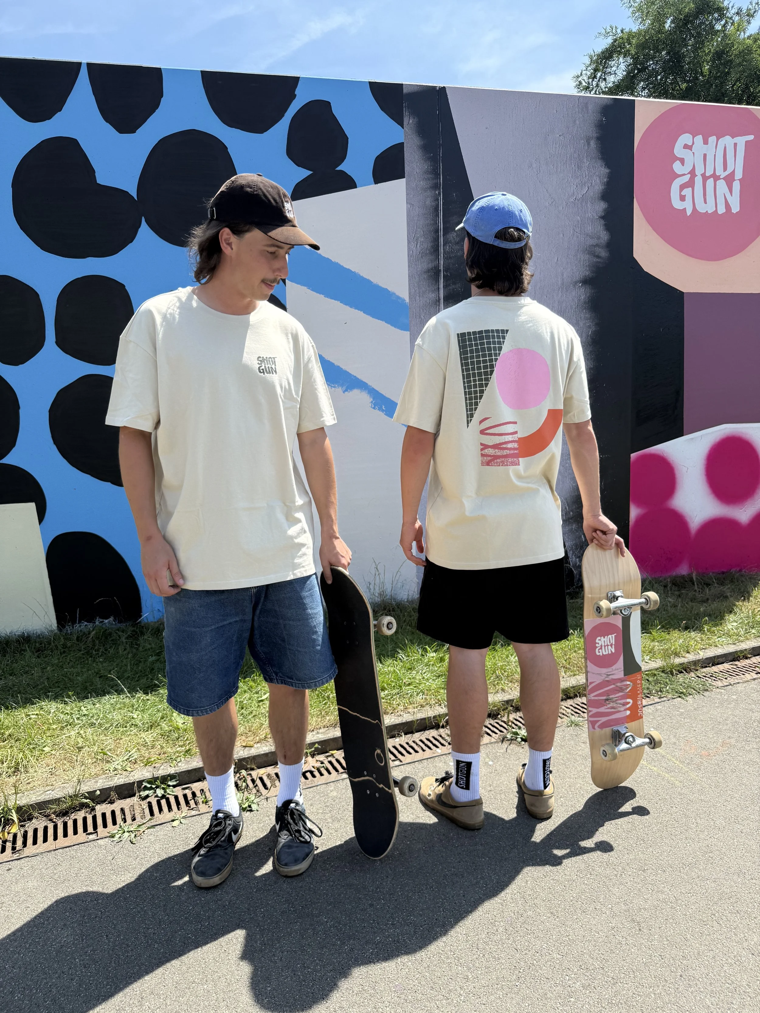 Zwei Skateboarder in „Shot Gun“-T-Shirts und Caps stehen mit Skateboards vor einer farbenfrohen Graffitiwand.