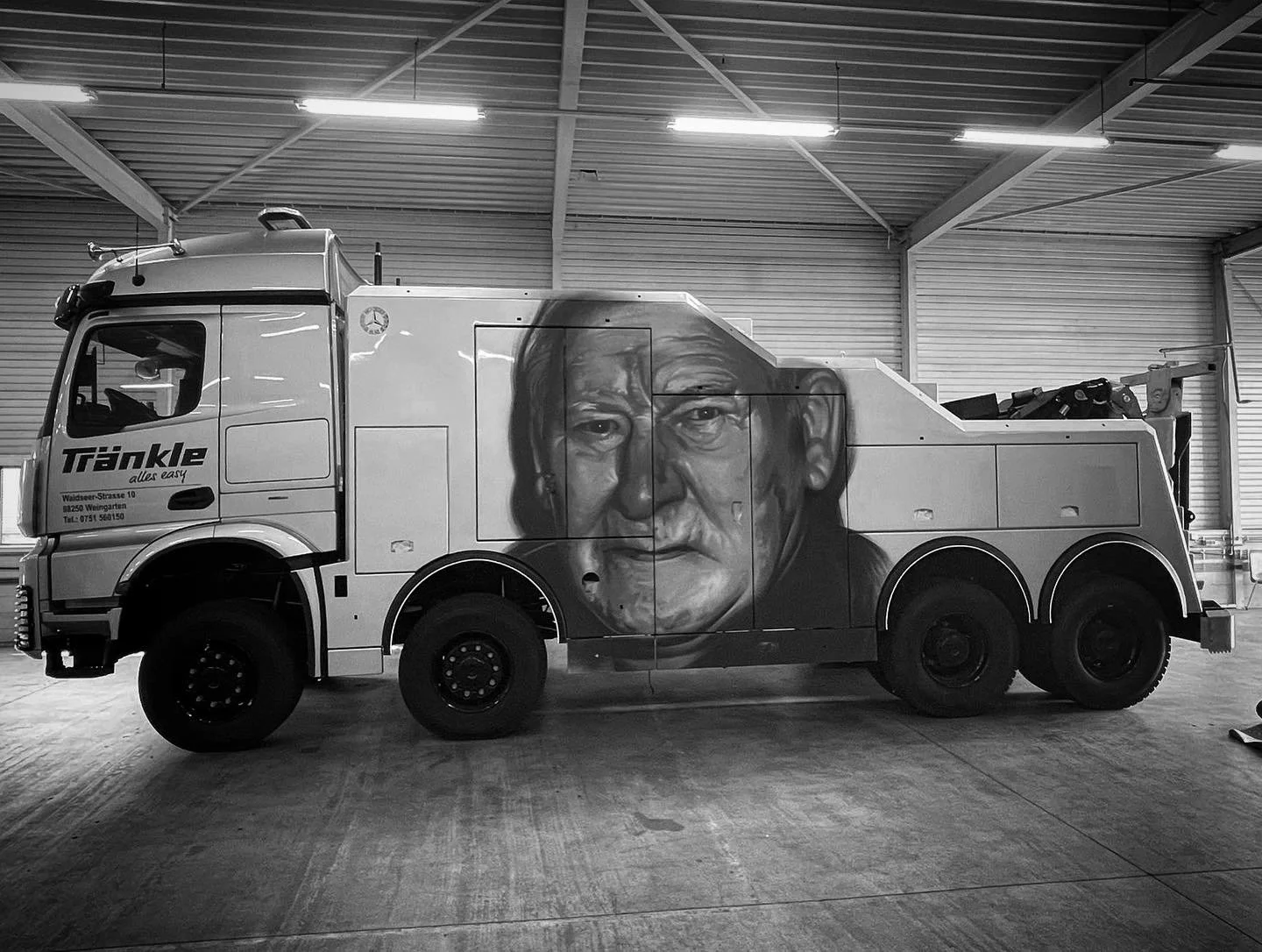 Ein mit Graffiti bemalter Abschleppwagen mit einem großen, realistischen Porträt eines älteren Mannes auf der Seite, im Inneren einer Lagerhalle.