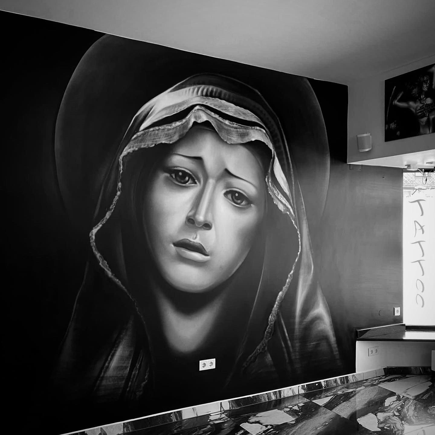 Schwarz-Weiß-Porträt von Maria mit traditioneller Kopfbedeckung, dargestellt auf einer Wand im Innenraum.