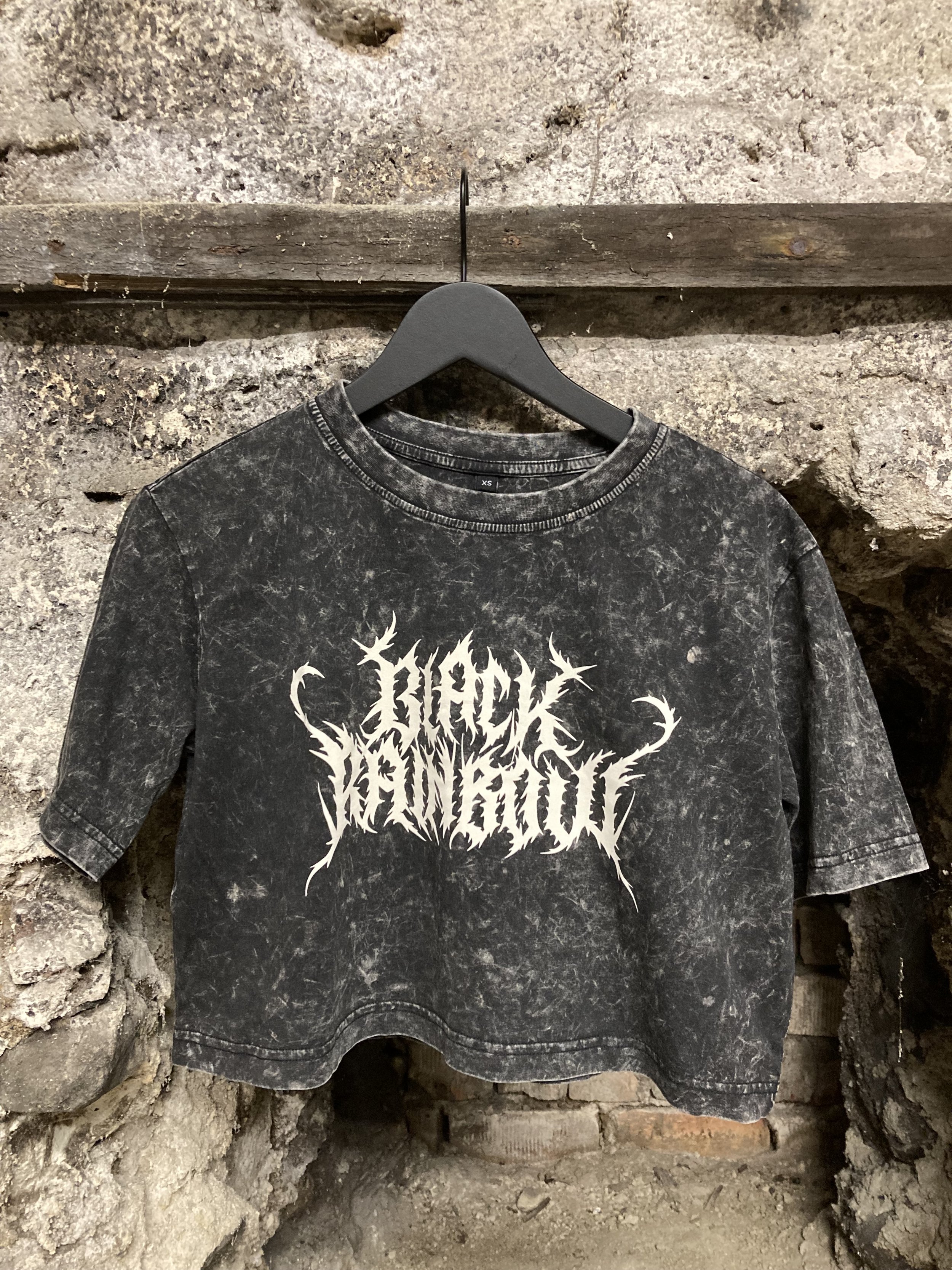Schwarzes, verwaschenes T-Shirt mit weißem Death-Metal-Logo auf einem Kleiderbügel vor einer rustikalen Steinwand.