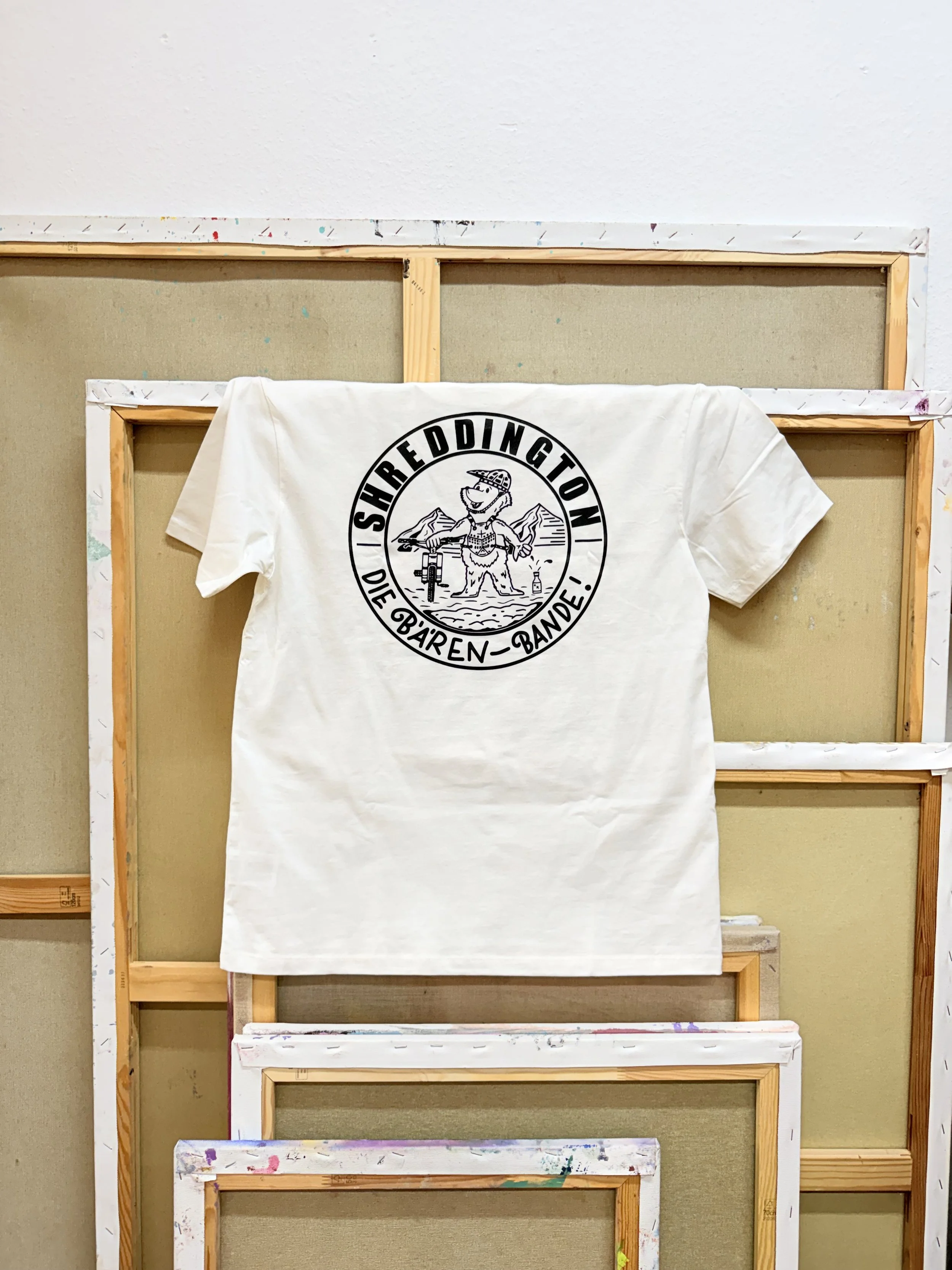 Weißes T-Shirt mit schwarzem Aufdruck „Shreddington Die Bären-Bande“: ein Bär mit Fahrrad vor Berglandschaft, vor mehreren leeren Bilderrahmen.