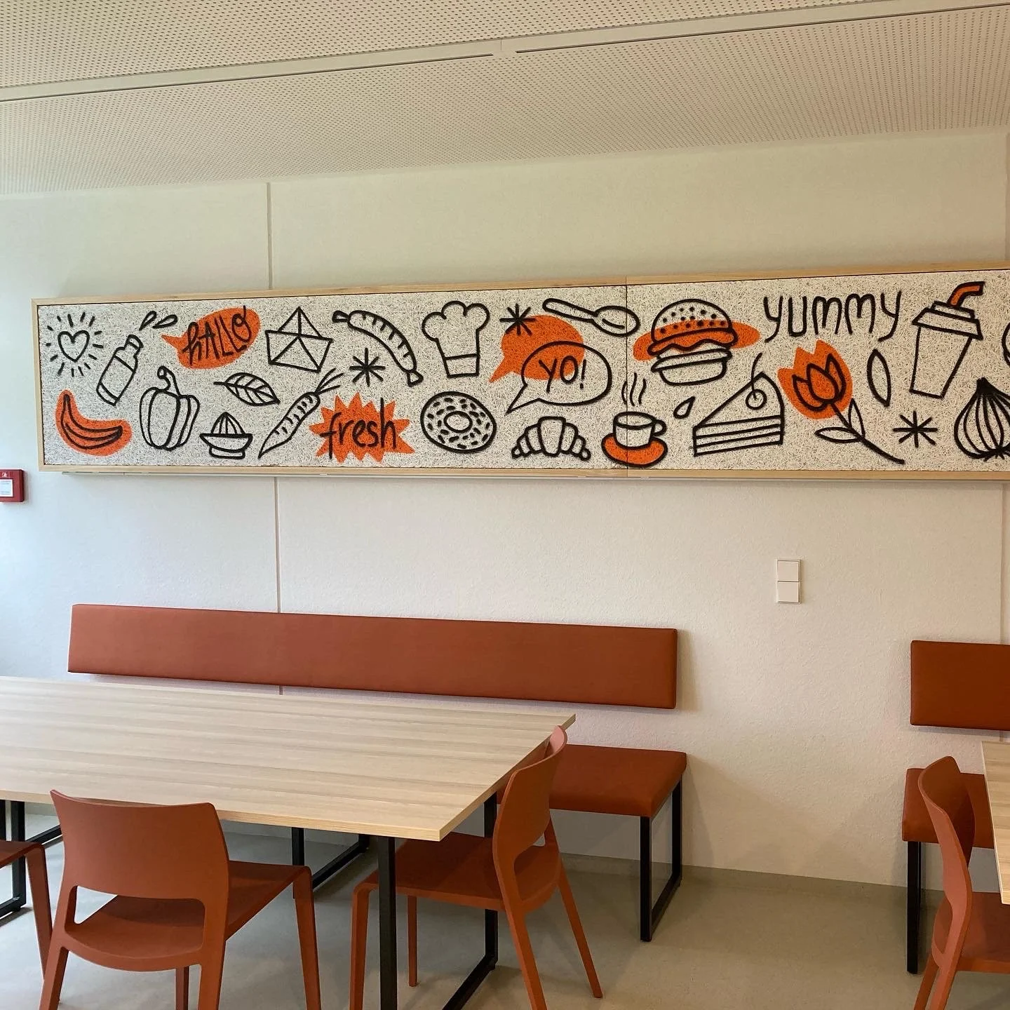 Eine Wand mit einer weißen Tafel, auf der handgezeichnete Lebensmittel und Wörter in Schwarz, Orange und Rot zu sehen sind, darunter Kaffetasse, Croissant, Burger, Löffel, Kräuter, belesene Worte wie 'hello', 'fresh', 'yo!', 'yummy'.