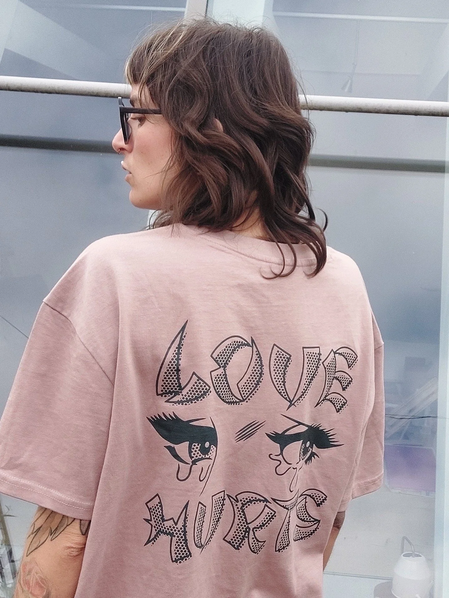 Person mit braunem, welligem Haar und schwarzer Brille trägt ein rosa T-Shirt mit auffälligem Cartoon-Print.