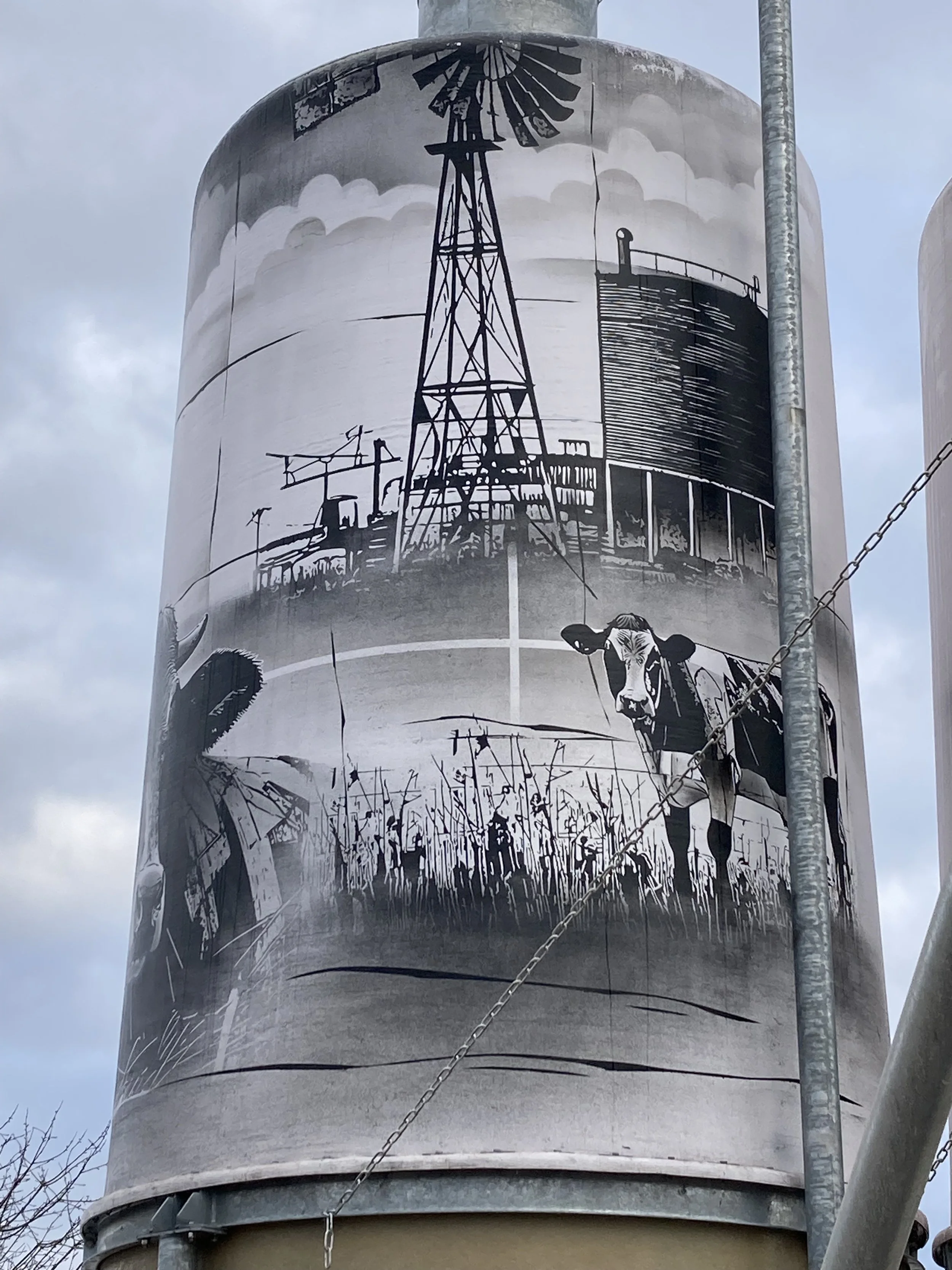 Schwarz-weiße Wandmalerei auf einem hohen zylindrischen Lagerbehälter, zeigt eine Windmühle, einen Kuhkopf und ländliche Landschaftsdetails.