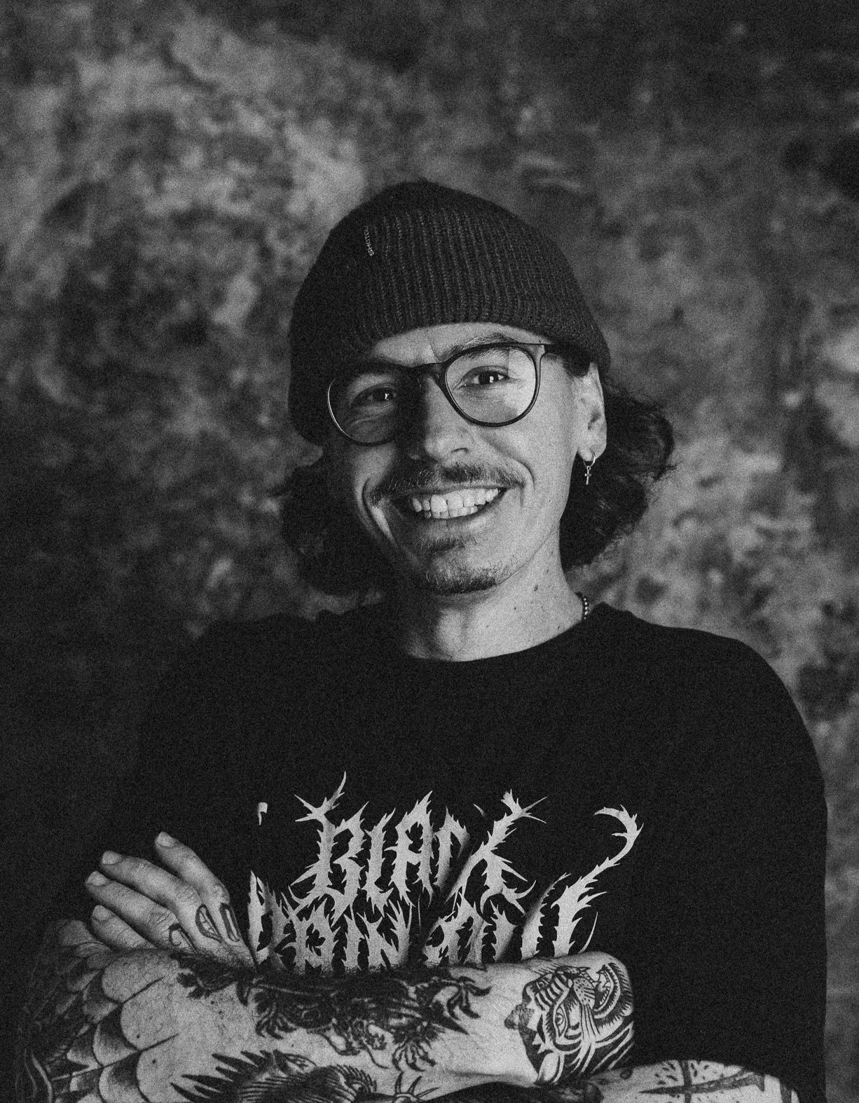 Schwarz-weiß-Foto eines lächelnden Mannes mit Brille, Beanie und Tattoos auf den Armen, vor einer steinigen Wand. Er trägt ein T-Shirt mit einem Metal-Band-Logo.