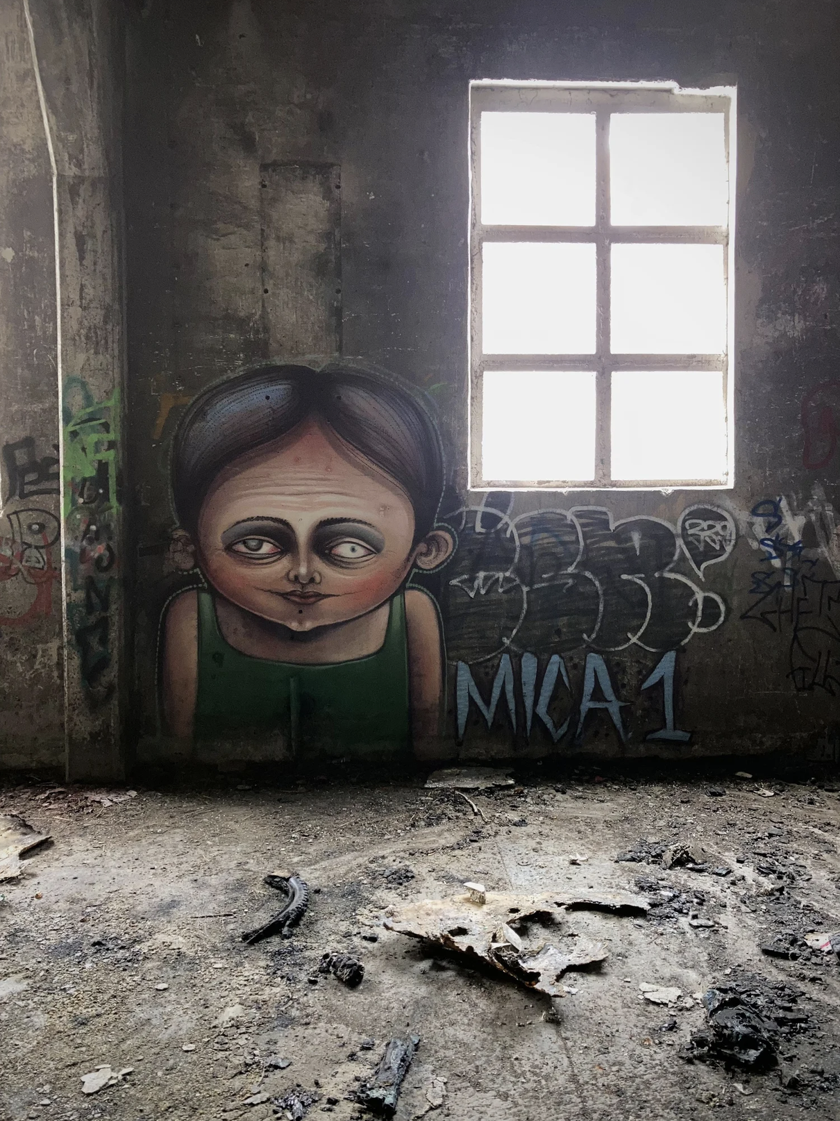 Graffiti eines kindlichen, cartoonartigen Gesichts mit braunen Haaren, grüner Kleidung und großen, makellosen Augen, in einer verlassenen, verfallenen Raum mit Graffiti an den Wänden und einem veralteten Fenster.