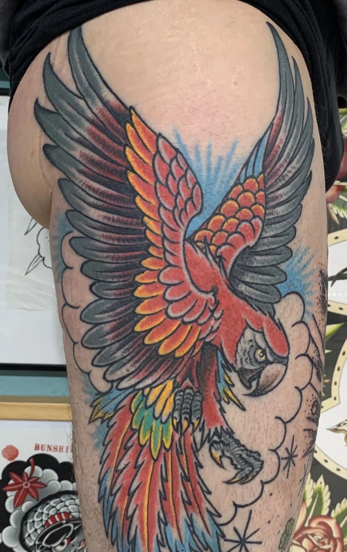 Buntes Tattoo eines fliegenden Papageien mit ausgebreiteten Flügeln und stilisierten Wolken auf einem Oberarm