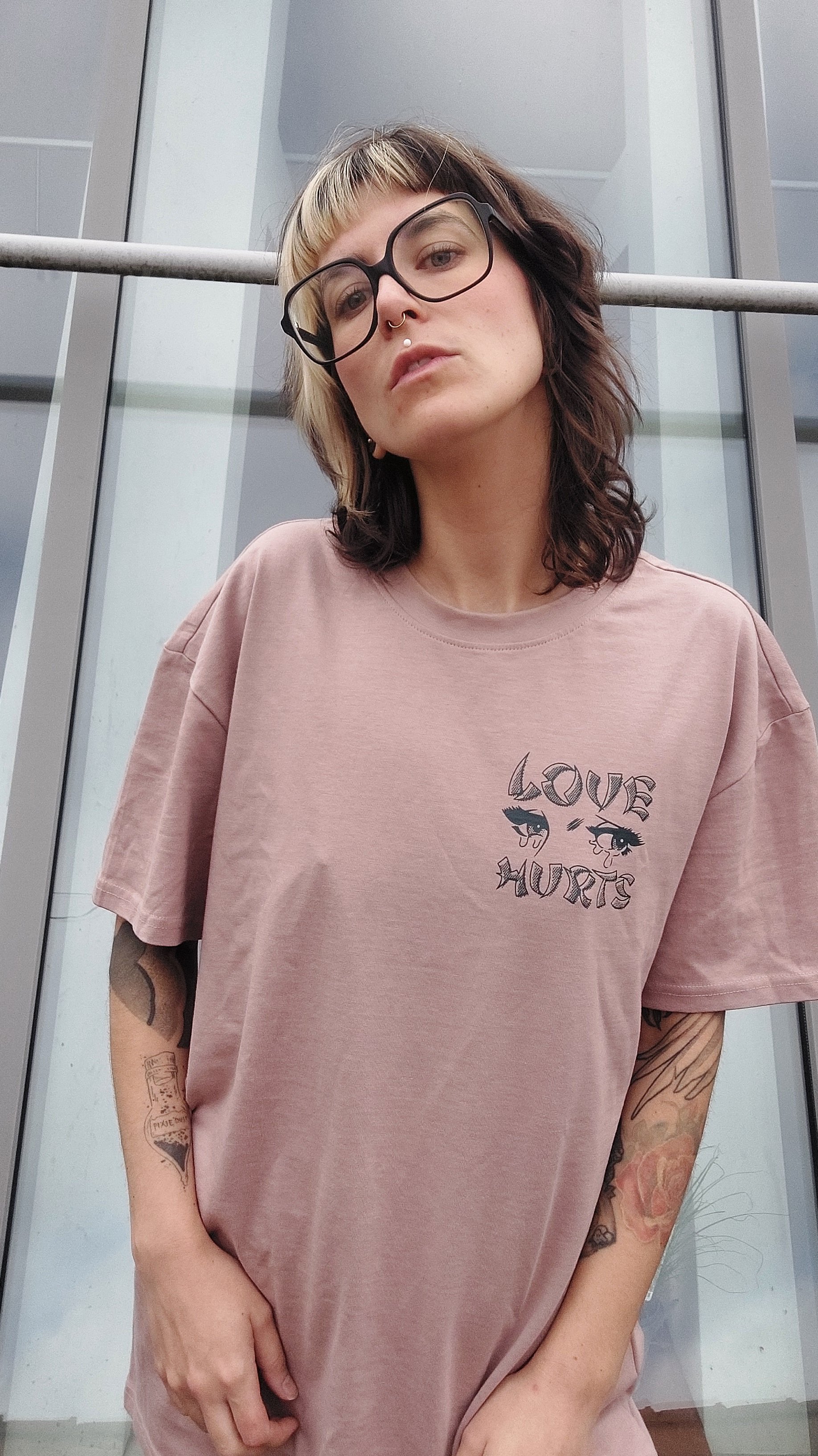 Porträt einer tätowierten Person mit zweifarbigen Haaren, großer schwarzer Brille, Nasen- und Lippenpiercing sowie rosa T-Shirt mit „LOVE HURTS“-Aufdruck vor einer Glasfassade.