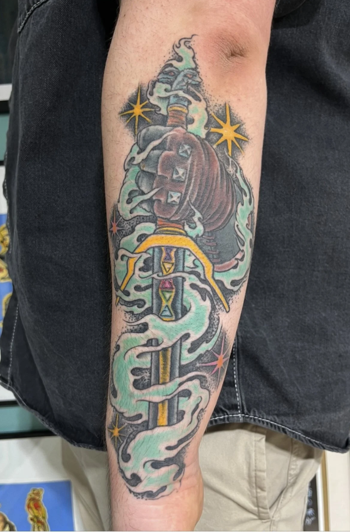 Farbenfrohes Tattoo am Unterarm mit einem grinsenden grünen Kobold, roten Umhang, goldenen Sternen und ornamentalen Rauchwolken.