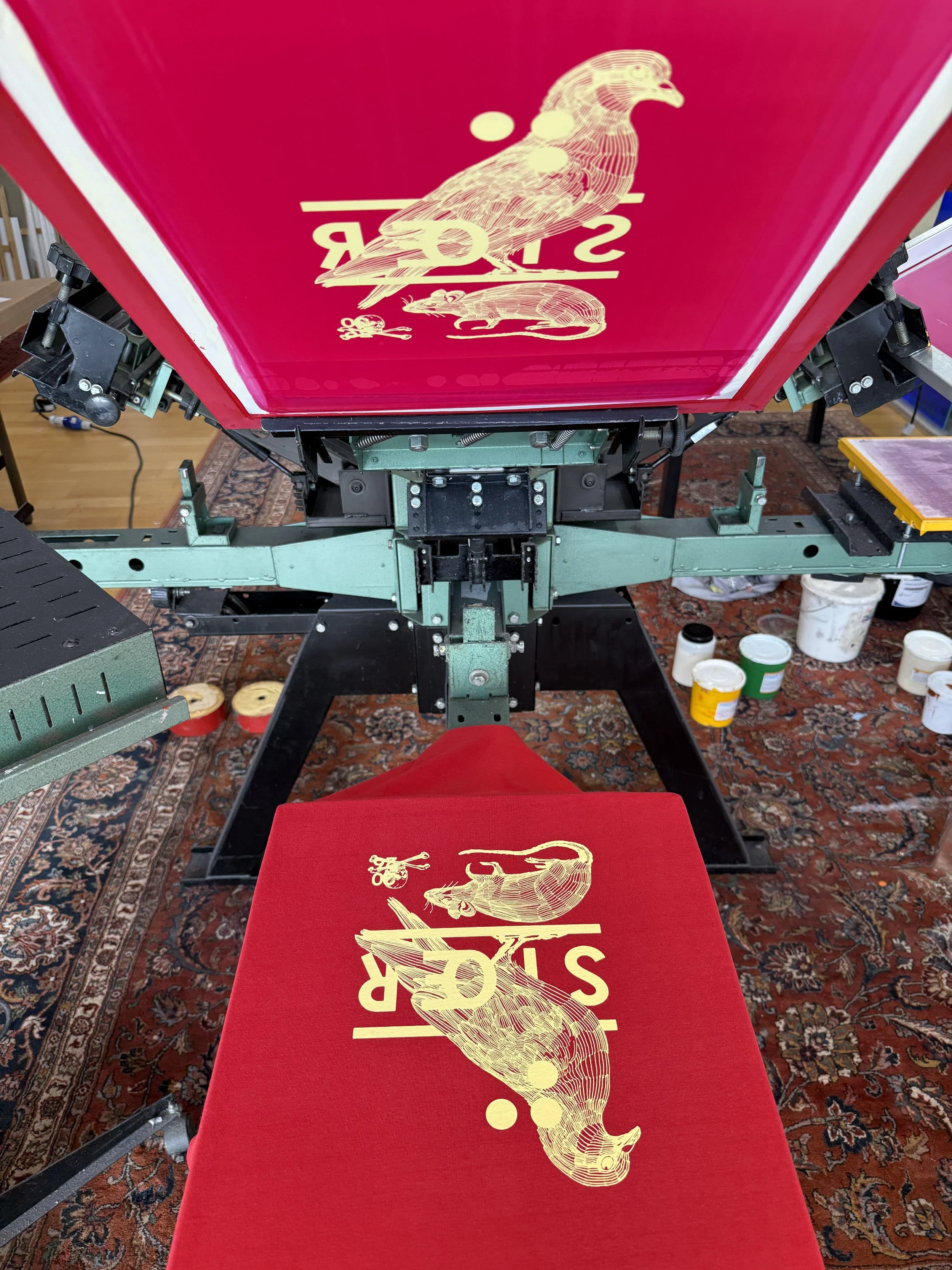 Rote Textilien mit goldenen Illustrationen von Vogel, Maus und Käfer in einer Siebdruckmaschine.