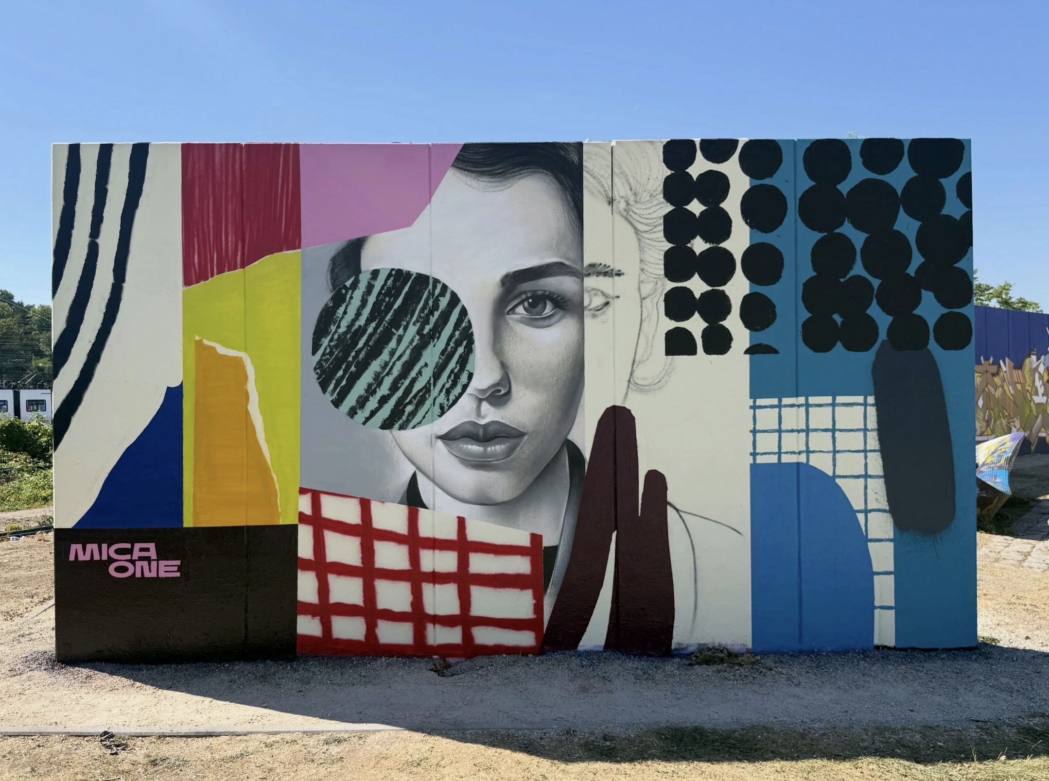 Graffiti-Mural mit einer schwarzen und weißen Porträtzeichnung eines weiblichen Gesichts, ergänzt durch bunte abstrakte und geometrische Muster in Gelb, Rot, Pink, Blau und Schwarz. Das Mural befindet sich an einer großen Wand außerhalb eines Gebäude