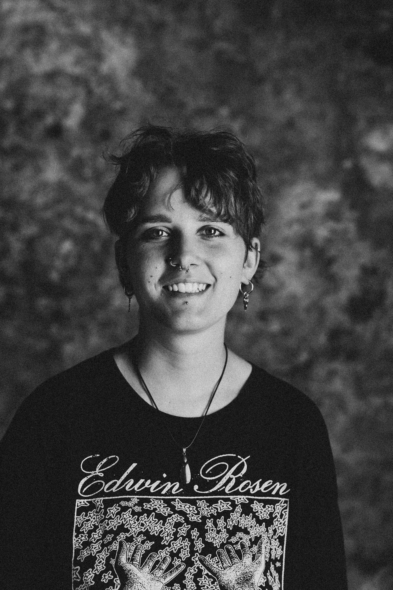 Schwarz-weiß Porträt einer jungen Frau mit kurzen, gewellten Haaren, mehreren Piercings im Gesicht, Ohrringen, Halskette, lächelnd, trägt ein T-Shirt mit dem Text 'Edwin Rosen' und einer Illustration von zwei Händen mit Sternen.