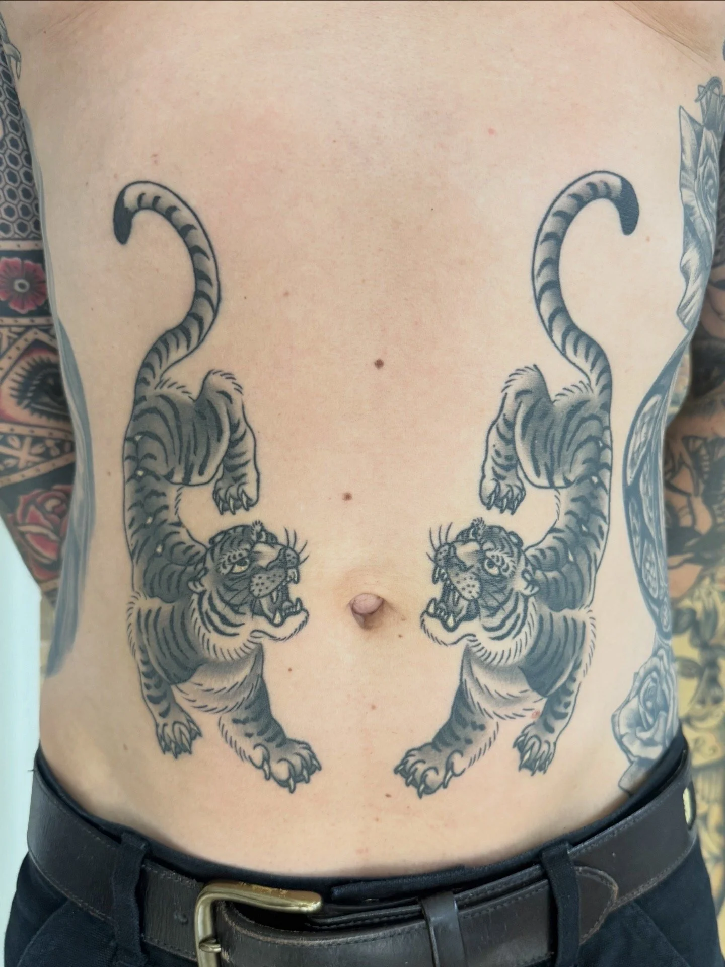 Nahaufnahme eines tätowierten Bauchs mit zwei spiegelbildlich angeordneten Tigern und dekorativen Tattoos an den Seiten.