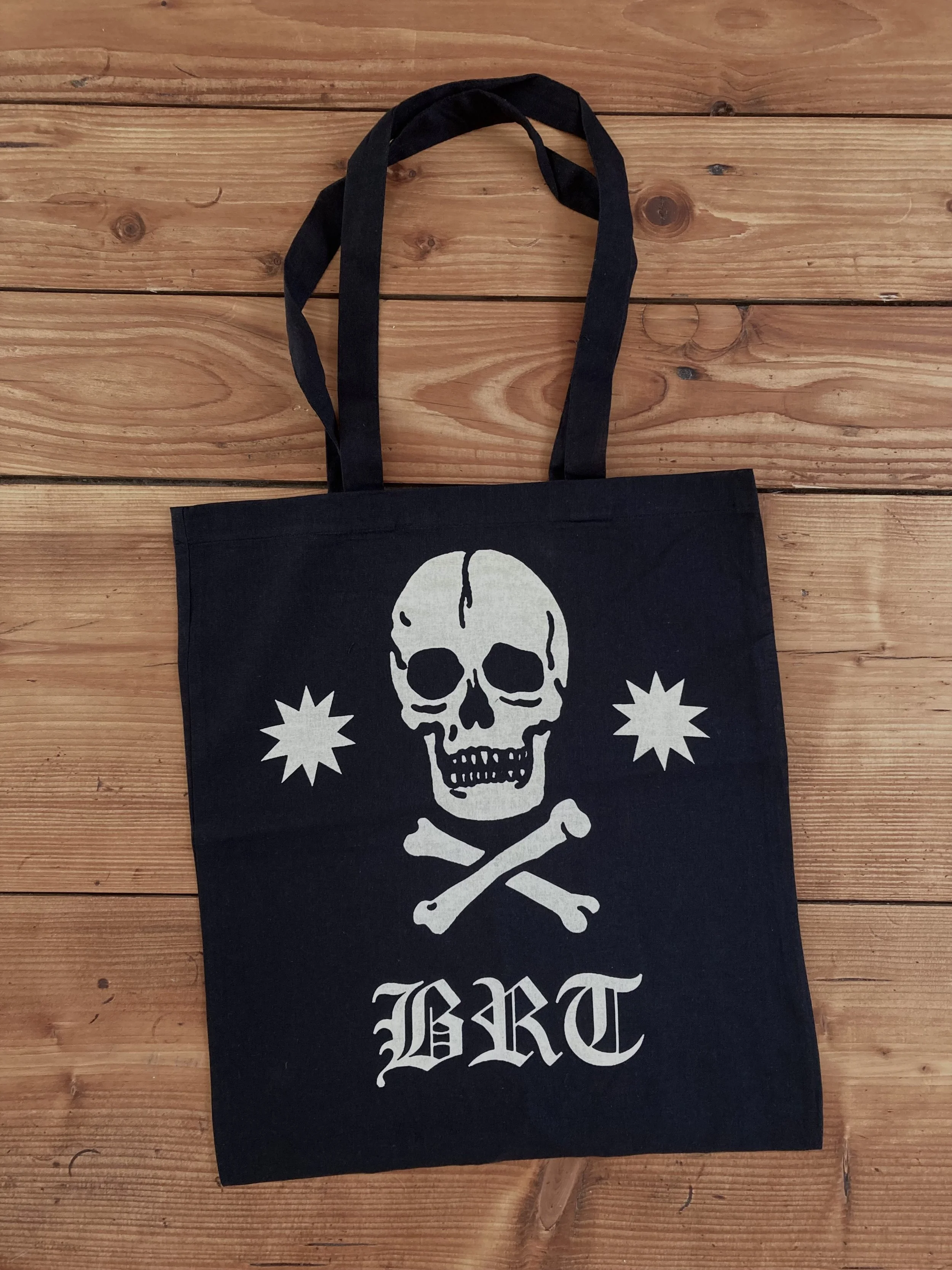 Schwarze Stofftasche mit weißem Totenkopf, gekreuzten Knochen, Sternen und dem Schriftzug „BRT“ auf einem Holztisch.