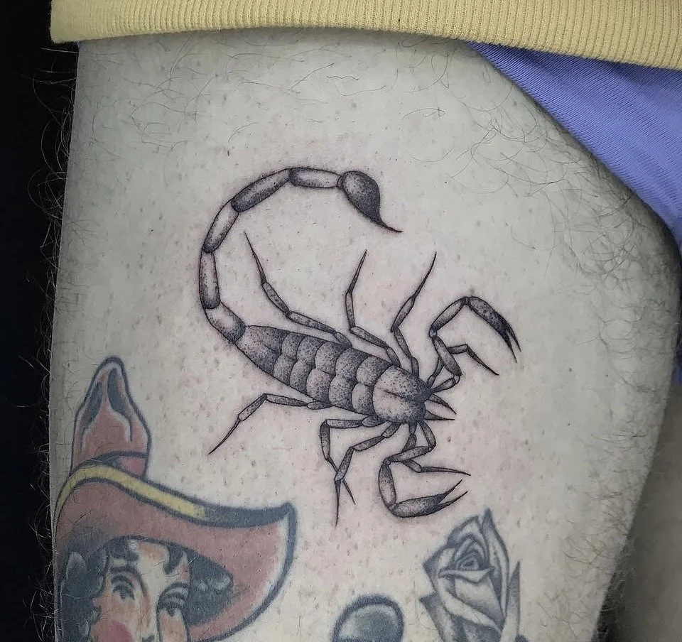 Detailaufnahme eines tätowierten Skorpions auf behaarter Haut, umgeben von weiteren bunten Tattoos.