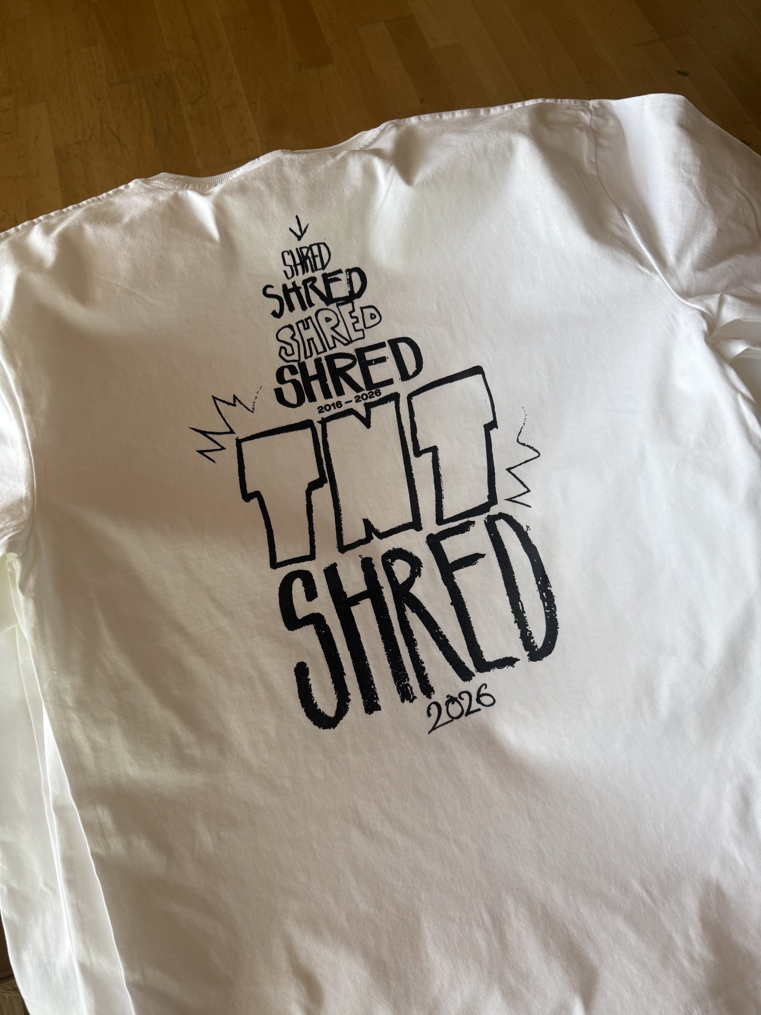 Nahaufnahme eines weißen T-Shirts mit schwarzem „Shred“-Aufdruck und der Jahreszahl 2026 auf einem Holzboden.