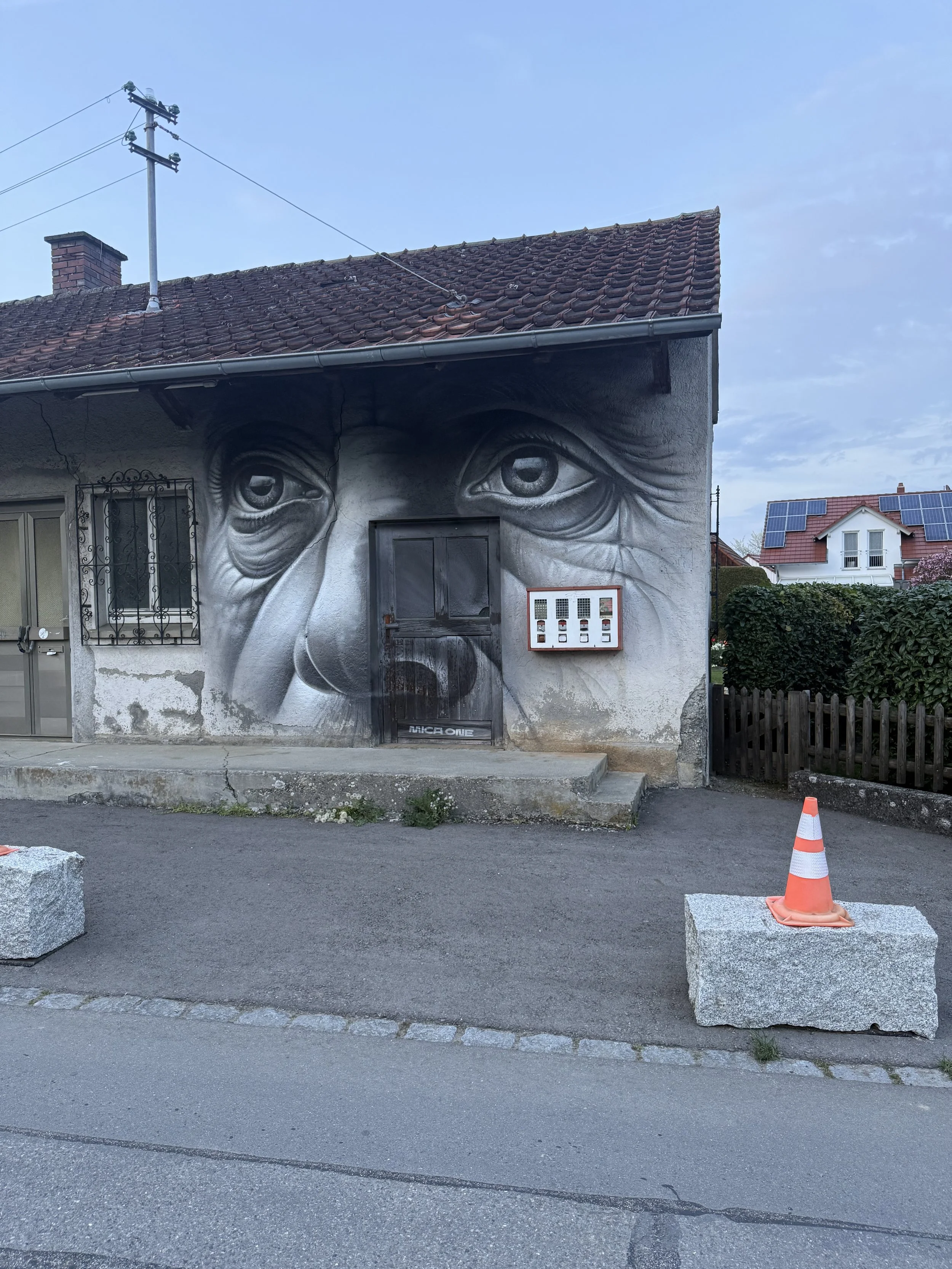 Eine Hauswand mit einem Wandgemälde eines menschlichen Gesichts, dessen Augen detailliert und realistisch gemalt sind. Es gibt eine Tür und ein Fenster mit Gitter, sowie eine Briefkastenanlage daneben. Im Vordergrund sind zwei große Steine und ein or