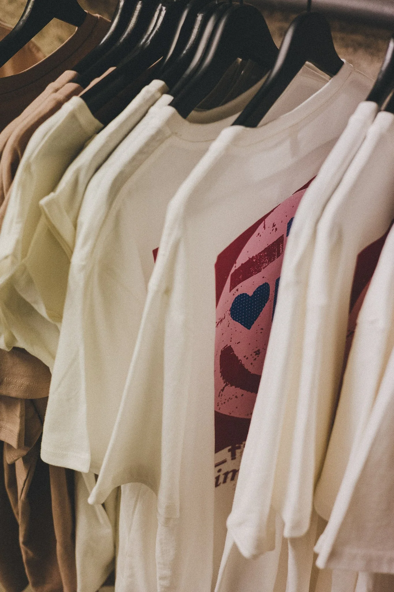 Nahaufnahme von T-Shirts auf Kleiderbügeln in verschiedenen Farben, darunter ein weißes Shirt mit rosafarbenem Aufdruck.