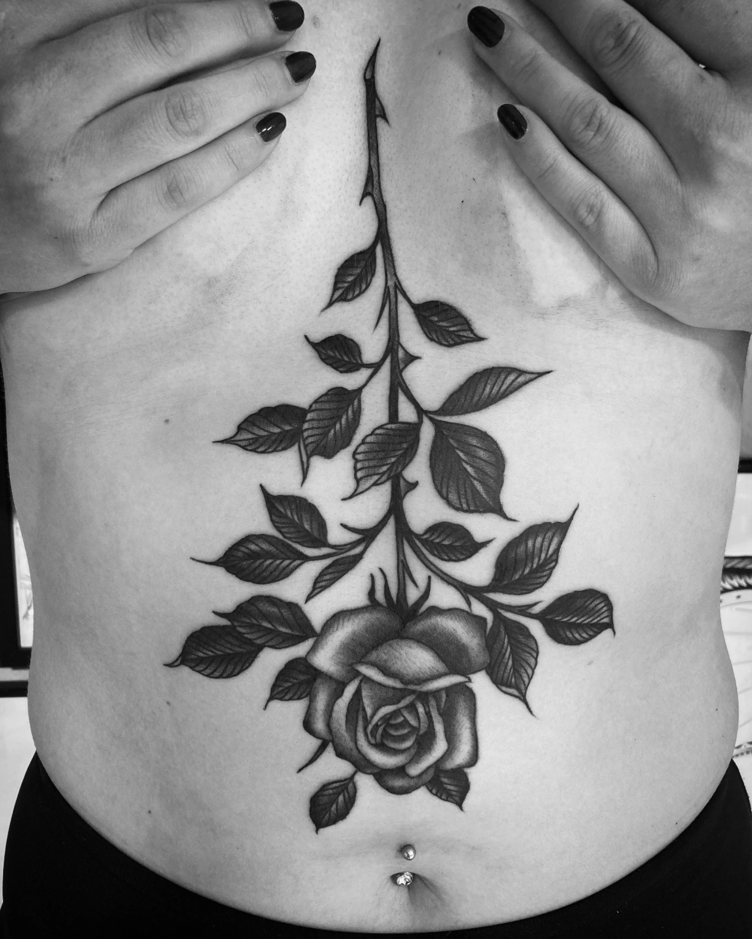 Schwarz-Weiß-Foto eines großen Rosen- und Blättertattoos auf dem Bauch, von Händen mit schwarzem Nagellack umrahmt.