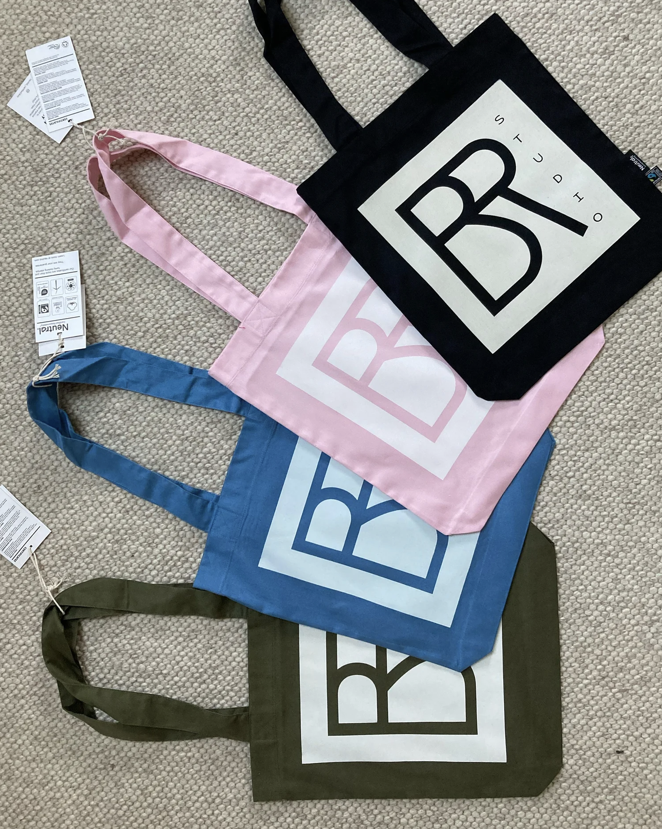Vier bedruckte Stofftaschen in Schwarz, Rosa, Blau und Olivgrün mit dem Schriftzug „BR Studio“ auf einem Teppich.