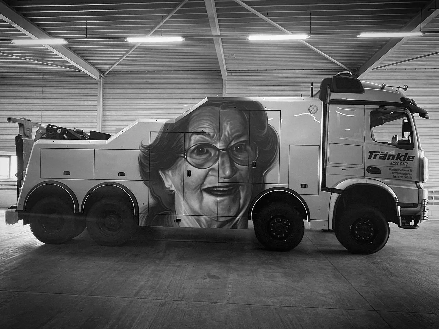 Ein LKW mit einem Gemälde einer älteren Frau mit Brille auf der Seite, aufgenommen in einer Lagerhalle im Schwarz-Weiß-Design.