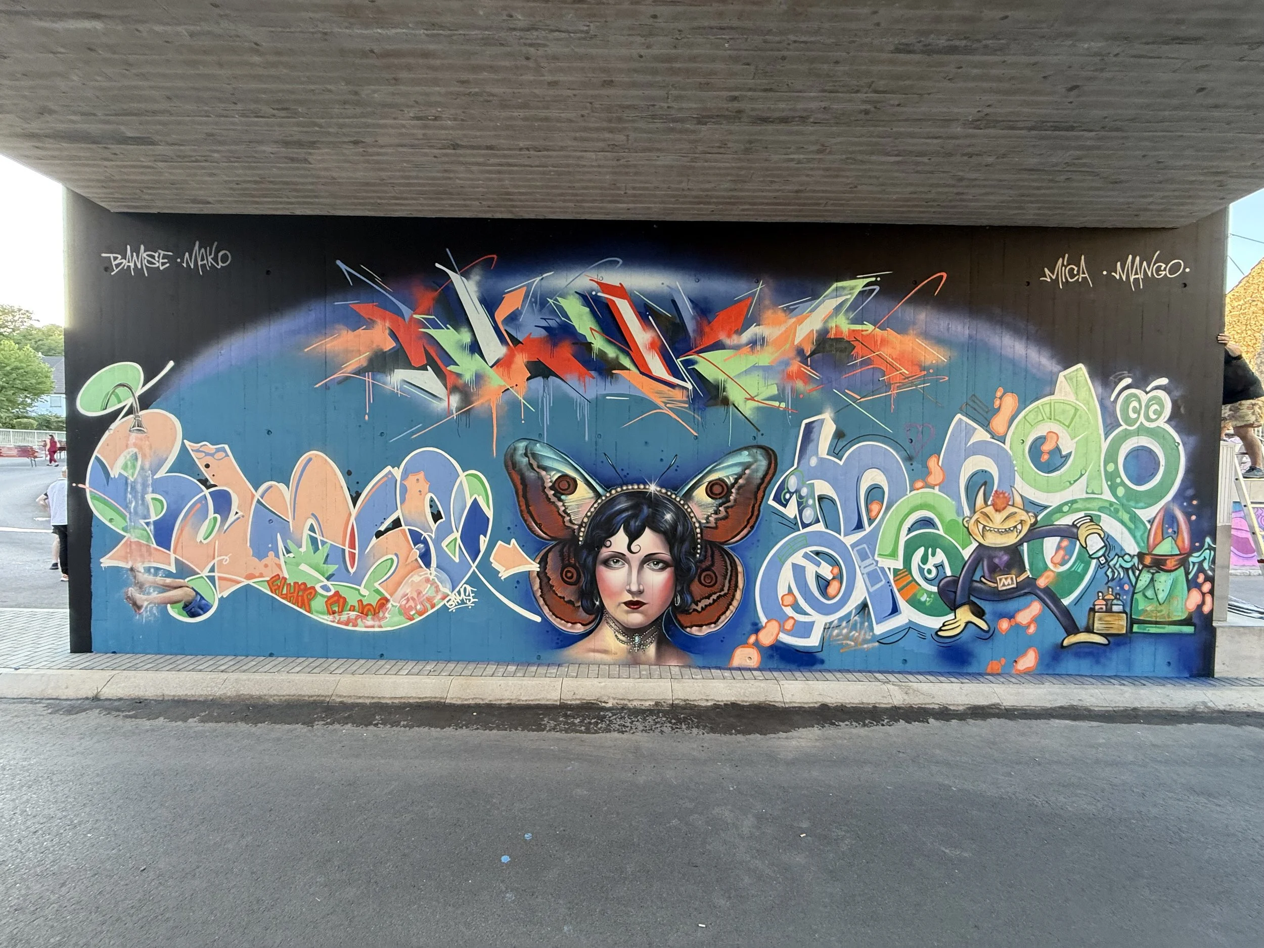 Graffiti an einer Wand unter einer Brücke, inklusive eines Porträts einer Frau mit schwarzen Haaren und auffälligem Make-up, bunten abstrakten Formen, einem Schmetterling und weiteren kreativen Elementen
