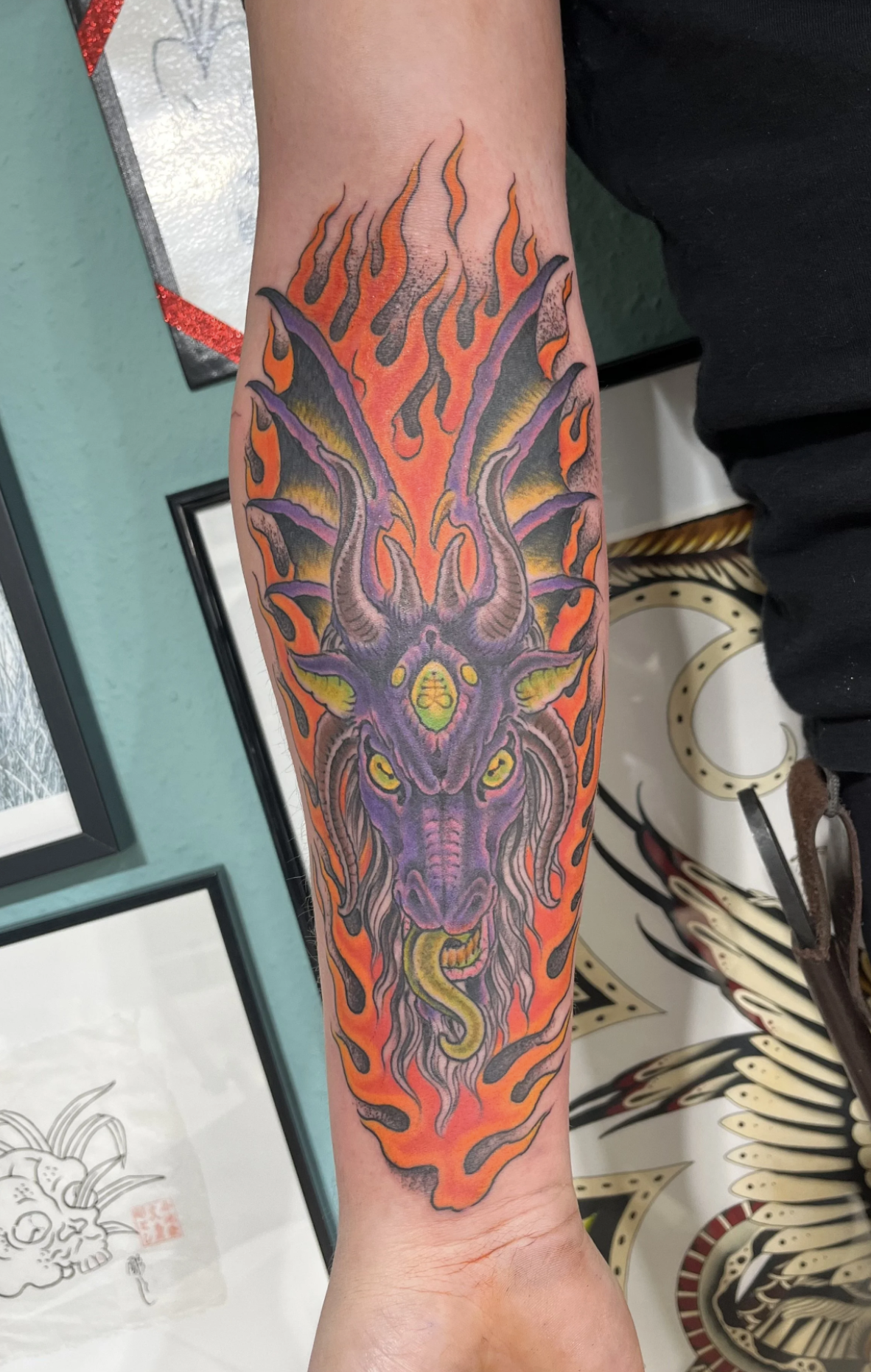 Farbenfrohes Unterarm-Tattoo mit dämonischem Drachen- oder Monsterkopf, grüner Zunge und Flammenmotiv in Orange, Violett und Schwarz.