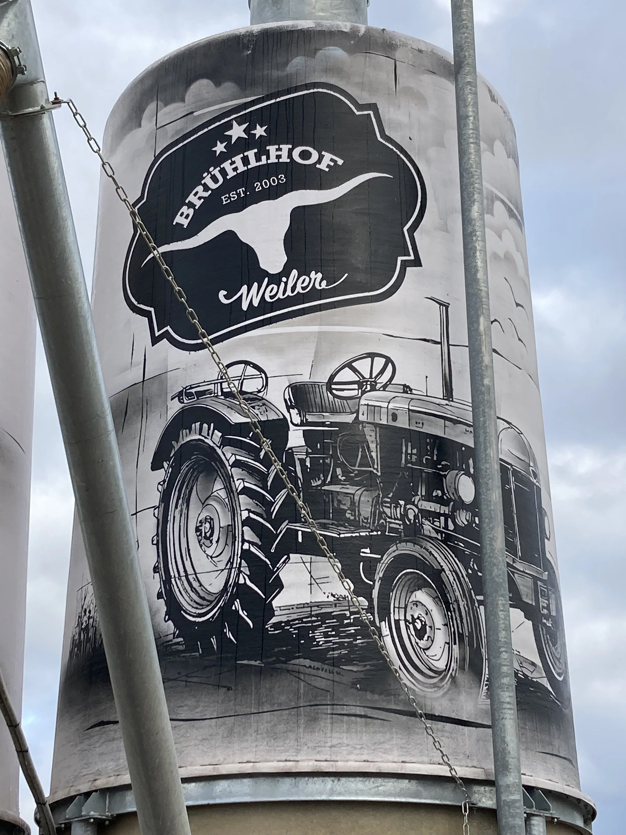 Ein Wasserbehälter mit einem Graffiti-Bild eines Traktors und einem Logo der Brauerei Brülhof Weiler, umgeben von Gerüsten und Kabeln.