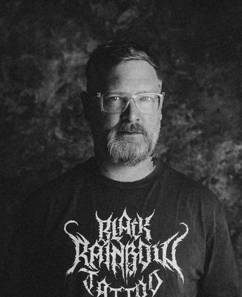 Schwarz-Weiß-Foto eines Mannes mit Brille, Bart und kurzen Haaren, trägt ein T-Shirt mit Metal-Logo. Hintergrund ist dunkel und unstrukturiert.