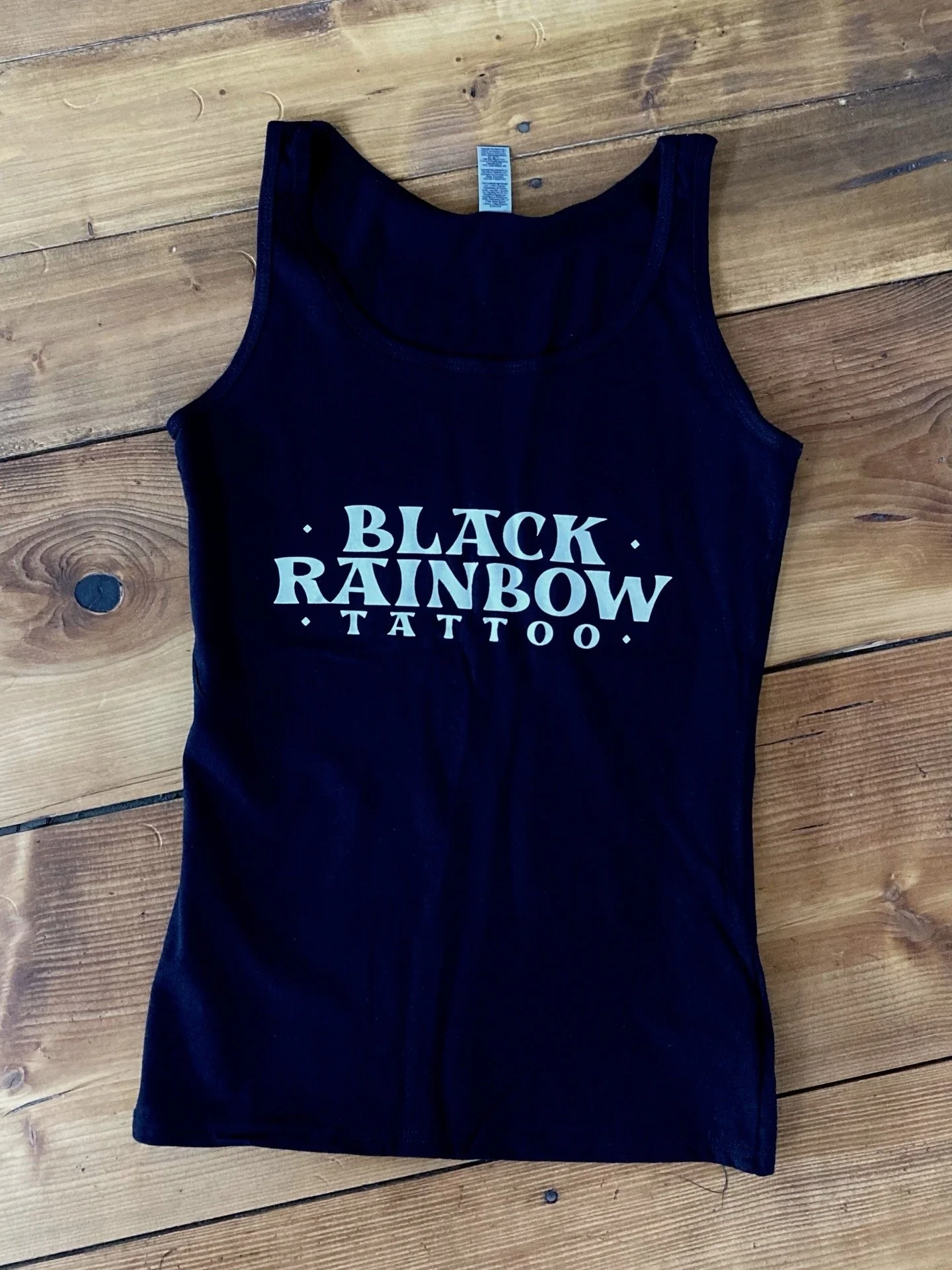 Schwarzes Tanktop mit weißem Aufdruck „BLACK RAINBOW TATTOO“ auf einem Holzboden.