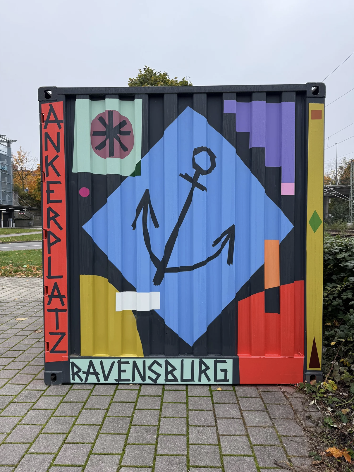 Das Bild zeigt eine bemalte Containerwand mit einem Anker in der Mitte, umgeben von bunten geometrischen Formen und Text. Rechts steht "RAVENSBURG" und links "DANKERPRÄSENT".