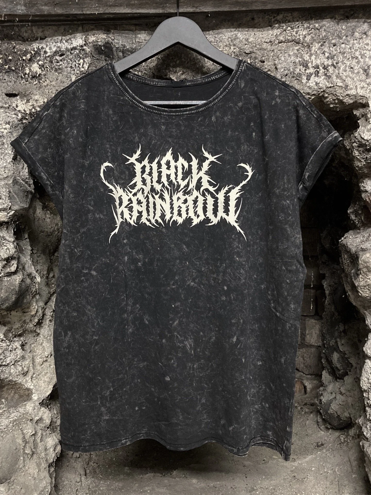 Schwarzes Vintage-T-Shirt mit weißem Death-Metal-Schriftzug auf einem Kleiderbügel vor einer Steinmauer