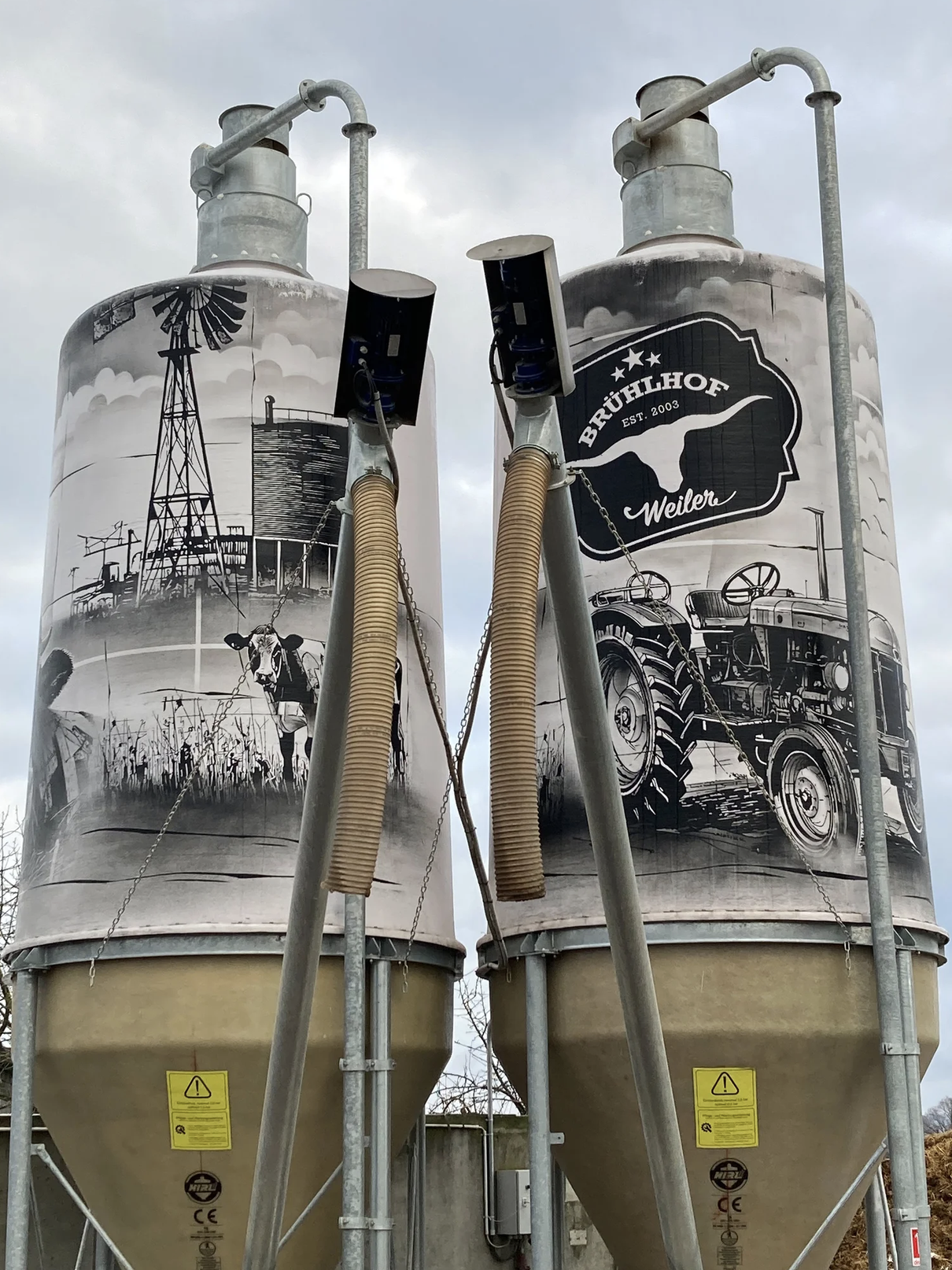 Zwei große Silos mit landwirtschaftlicher Dekoration, darunter eine Kuh, eine Windmühle, ein Traktor, ein Heuballen und eine Scheune, mit einem Firmenlogo "Brühlhof Weiler".