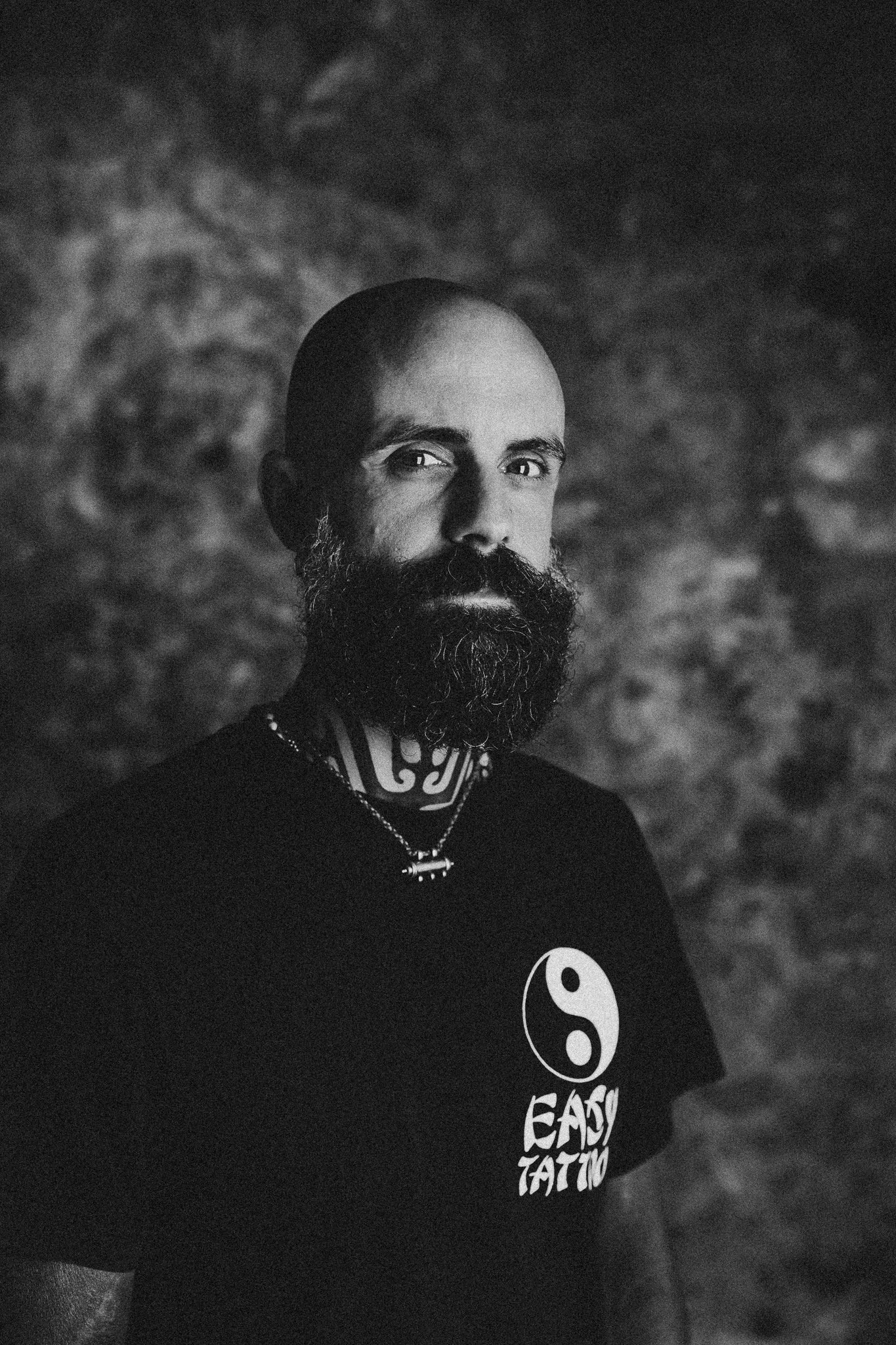 Schwarz-weiß-Foto eines Mannes  Bart, der eine T-Shirt mit der Aufschrift 'SO EASY TATTOO' trägt, auf einem dunklen, verschwommenen Hintergrund.