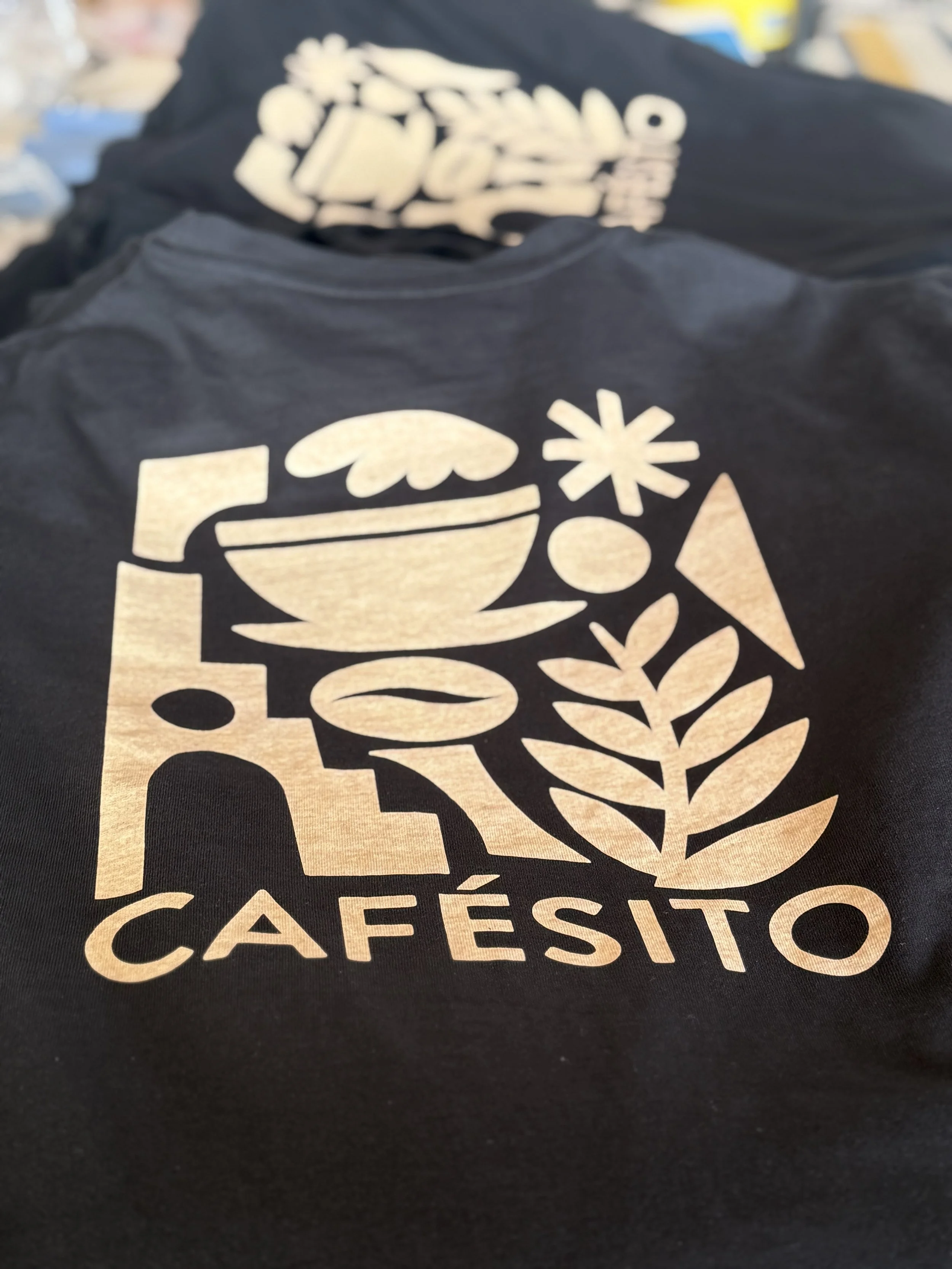 Nahaufnahme eines schwarzen T-Shirts mit goldener Grafik, Schriftzug „CAFESITO“ und stilisierten Kaffee- sowie Pflanzensymbolen.