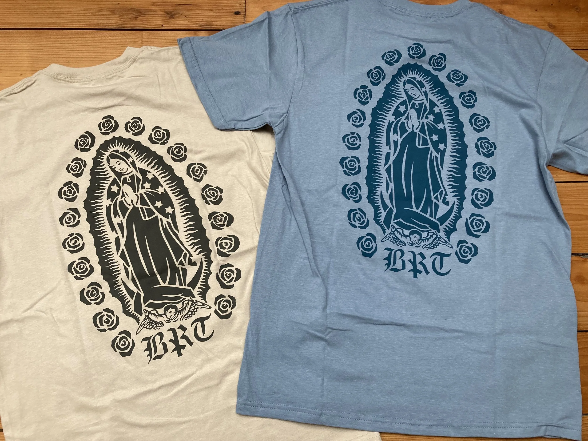 Zwei T-Shirts mit stilisiertem Marienbild, Rosenkranzmotiv und der Aufschrift „BRT“ auf einem Holztisch.