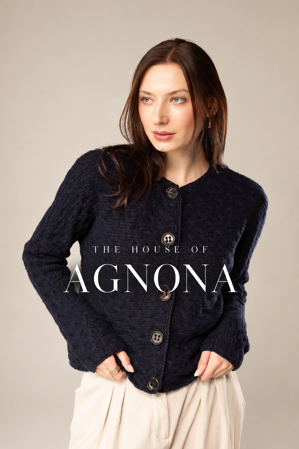 A woman with shoulder-length brown hair wearing a navy textured button-up cardigan and cream-colored pants, standing against a plain background with the text 'The House of Agnona' overlaid.