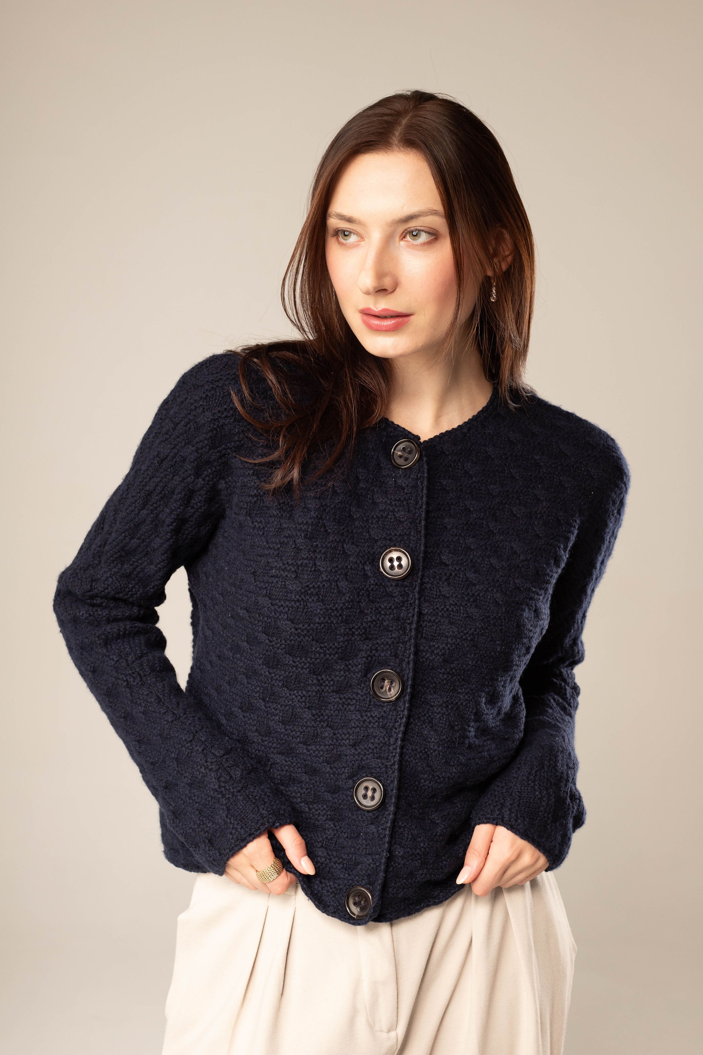 A woman with long dark hair wearing a navy blue textured button-up cardigan and cream-colored pleated skirt, standing against a plain beige background.