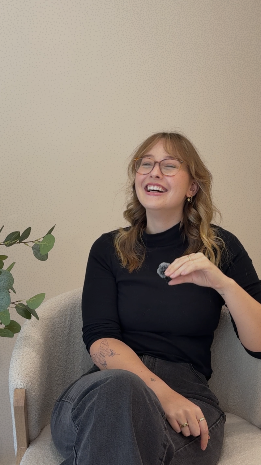 Smiling woman with glasses holding a small microphone while seated in an armchair beside a houseplant