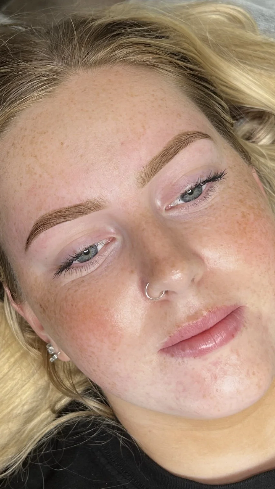 Close-up portrait of a freckled blonde woman with blue eyes, defined eyebrows, long eyelashes, and a silver septum ring