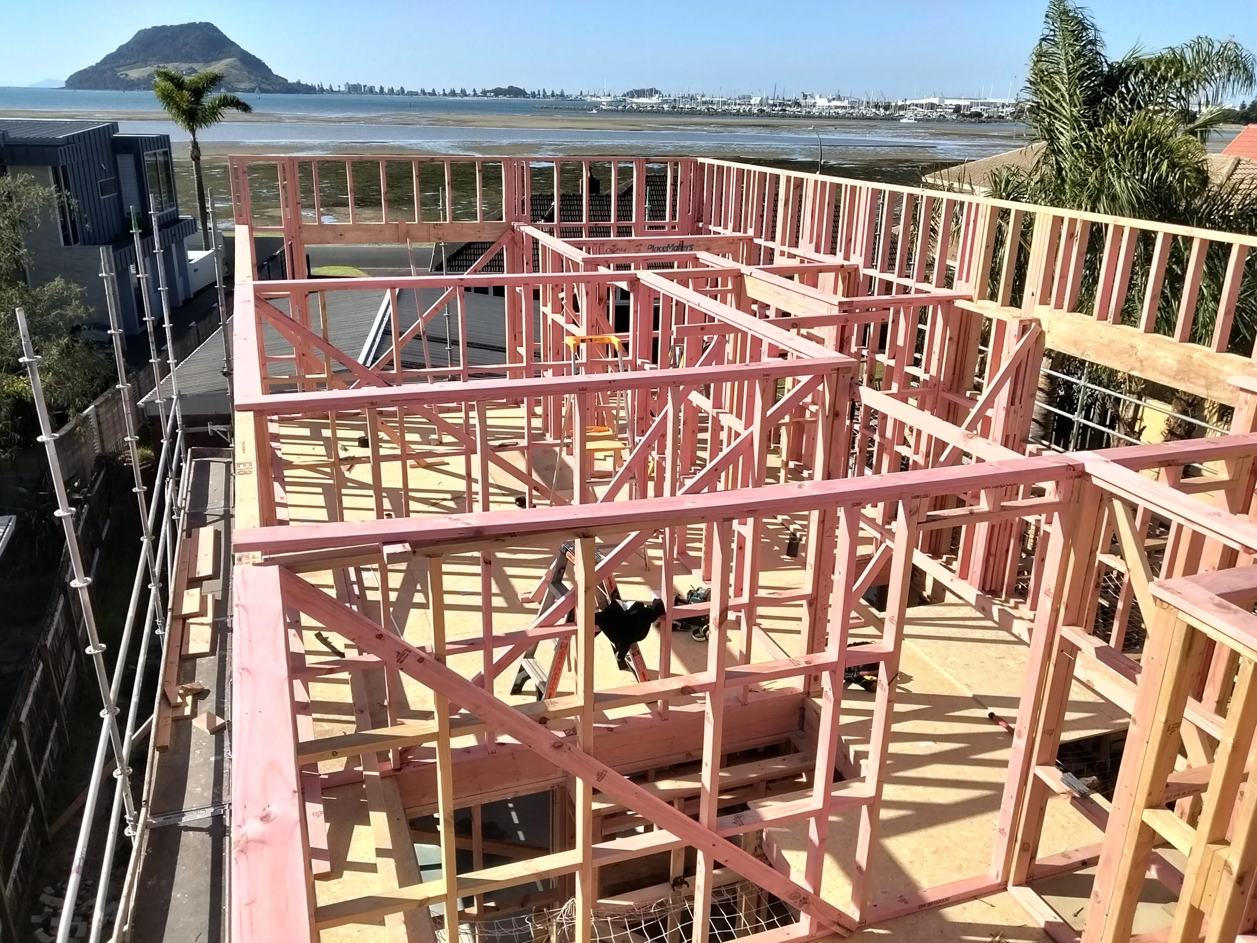 Construction of a wooden building with framing in progress overlooking a coastal landscape with water, boats, and hills in the background.