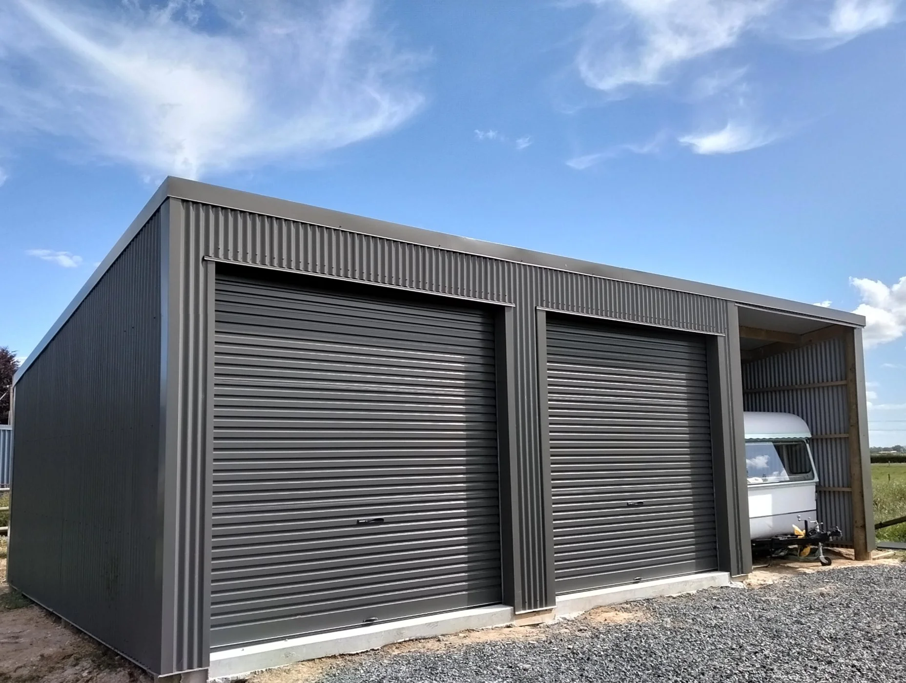 A metal building with two large gray rolled-up garage doors and a smaller open section on the right, with a white vehicle partially visible inside, on a gravel surface with a partly cloudy sky overhead.