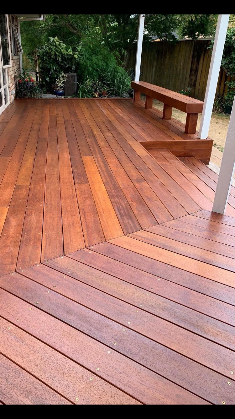 A wooden deck with reddish-brown planks, a built-in bench, and a garden with lush green plants and flowers in the background.