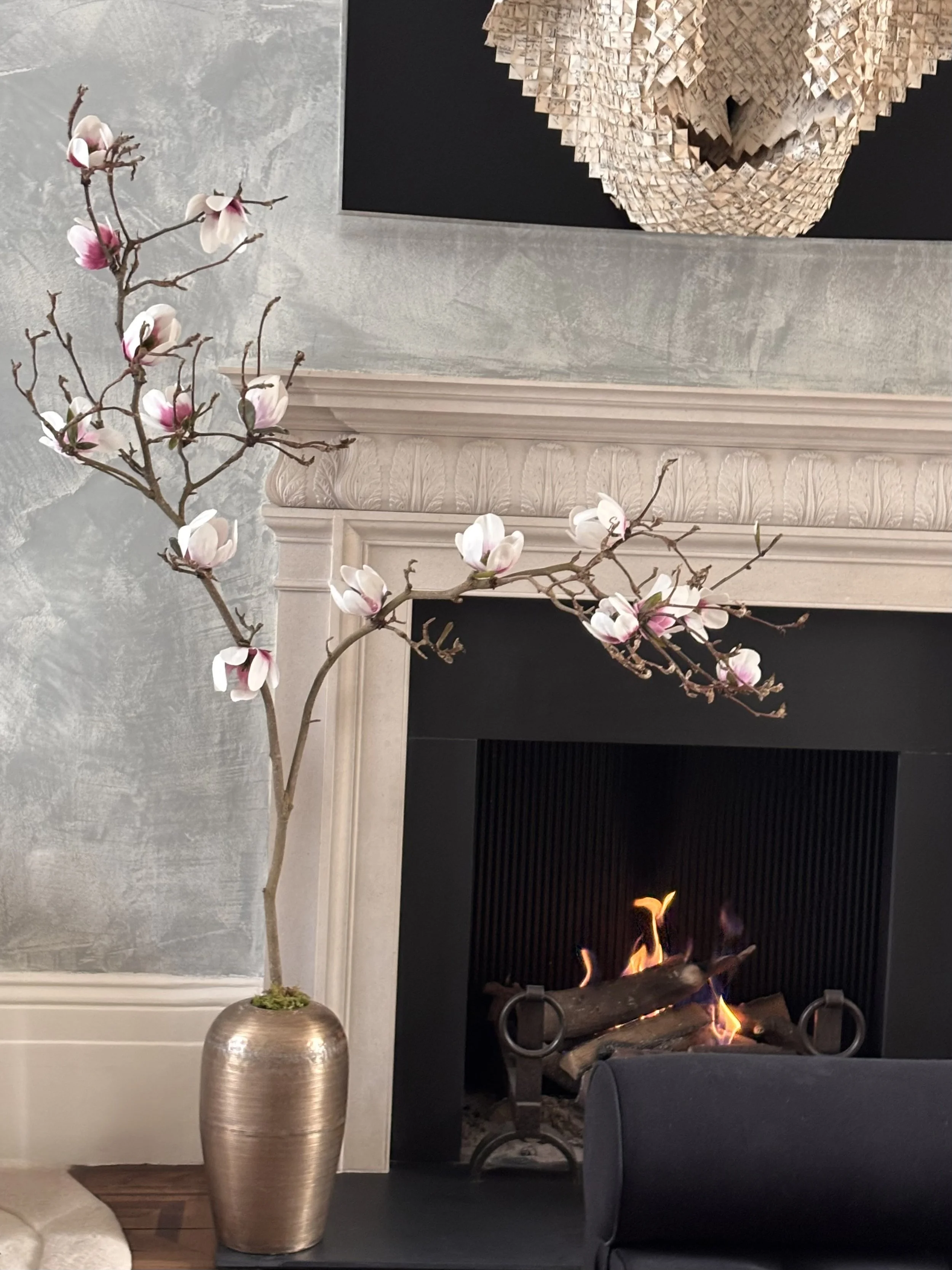 A decorative interior scene featuring a tall, metallic vase with pink and white magnolia flowers, a fireplace with a visible fire, a framed artwork with a textured heart shape on a black background, a wall with a two-tone design, and a plush beige area rug.