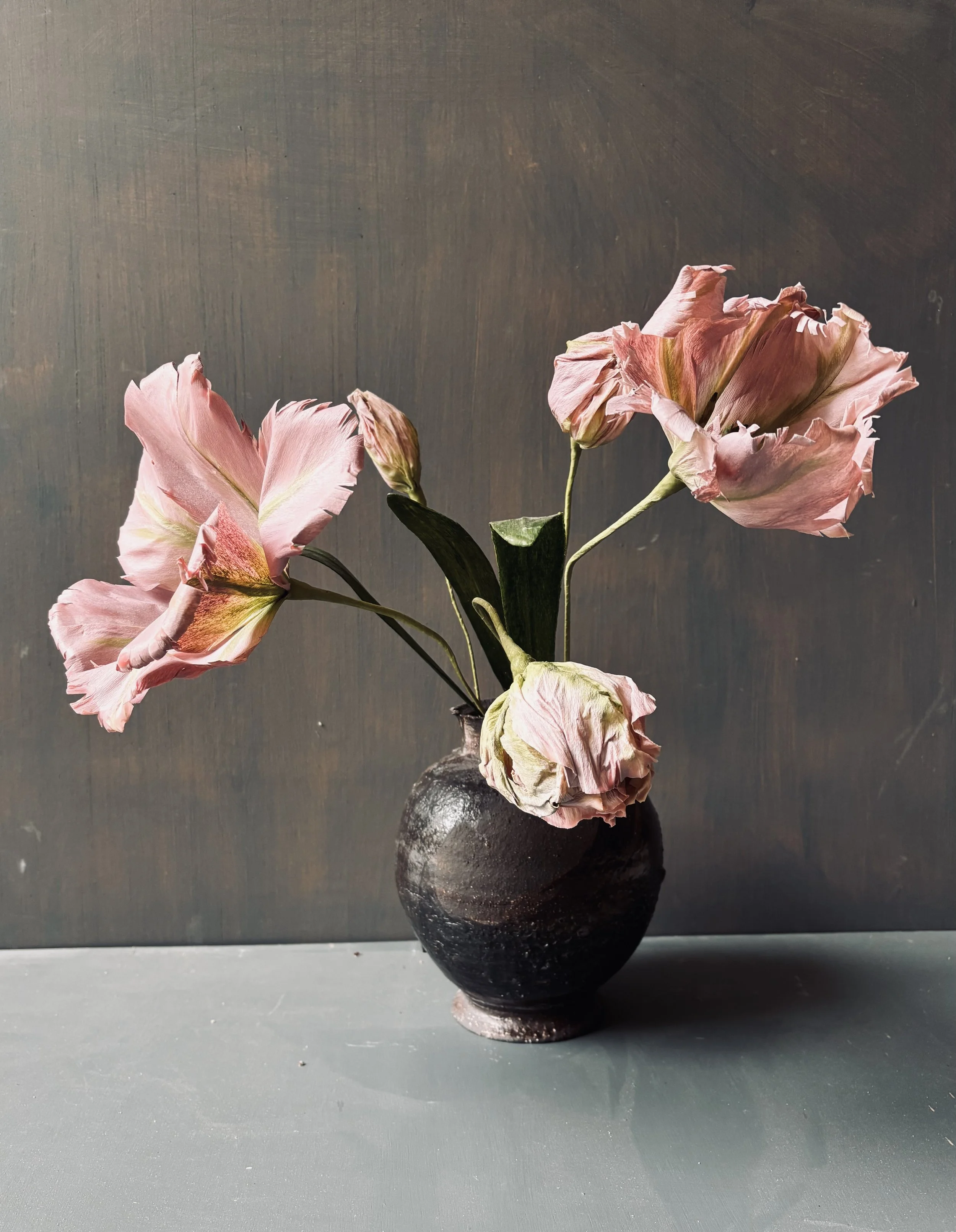 A small black vase with a rough texture holds wilted pinkish peony flowers with green stems and leaves, set against a dark textured background.